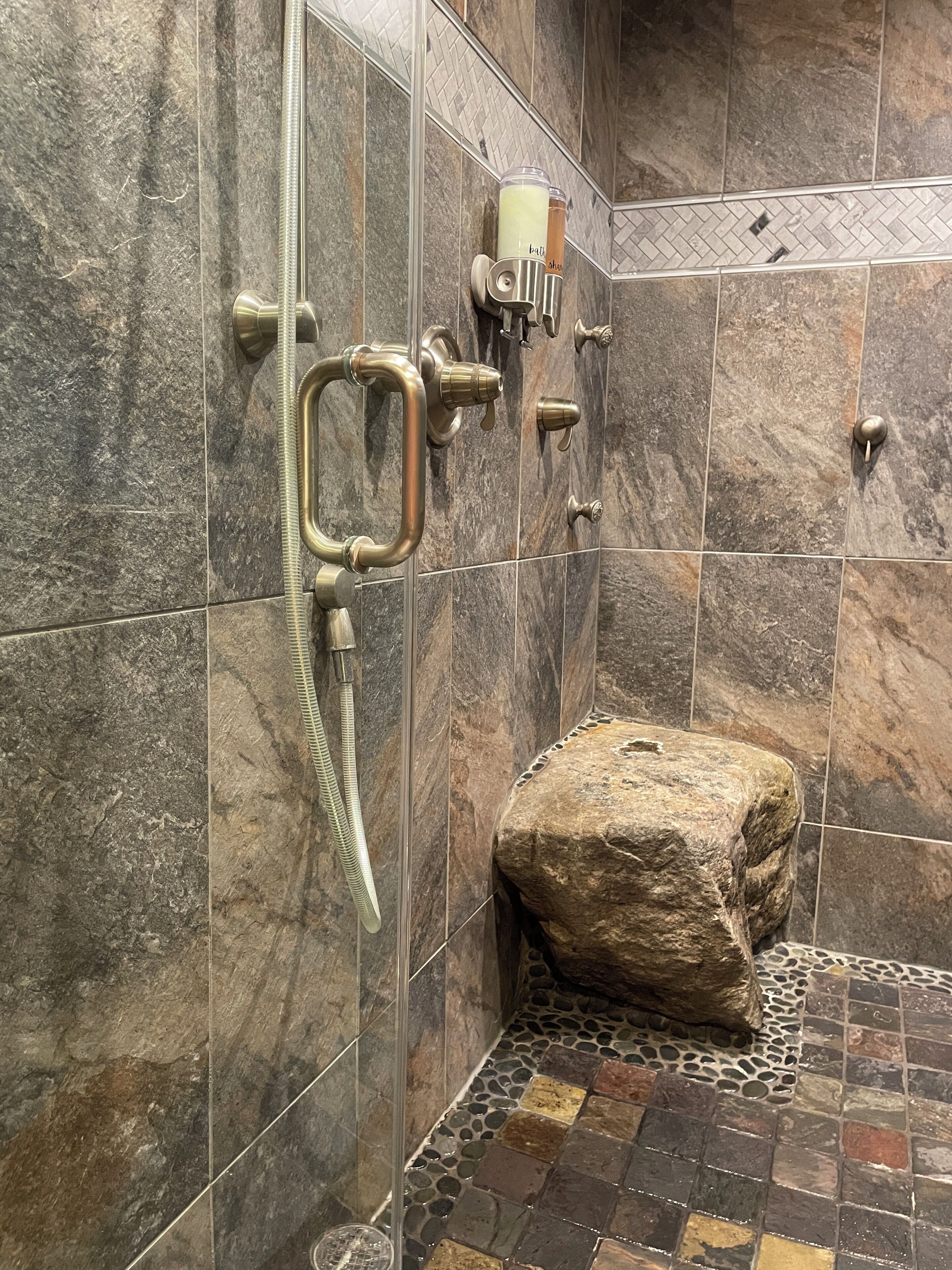 Un espacio de ducha con pared de piedra, cabezal de ducha, ducha de mano y una roca colocada en el suelo.