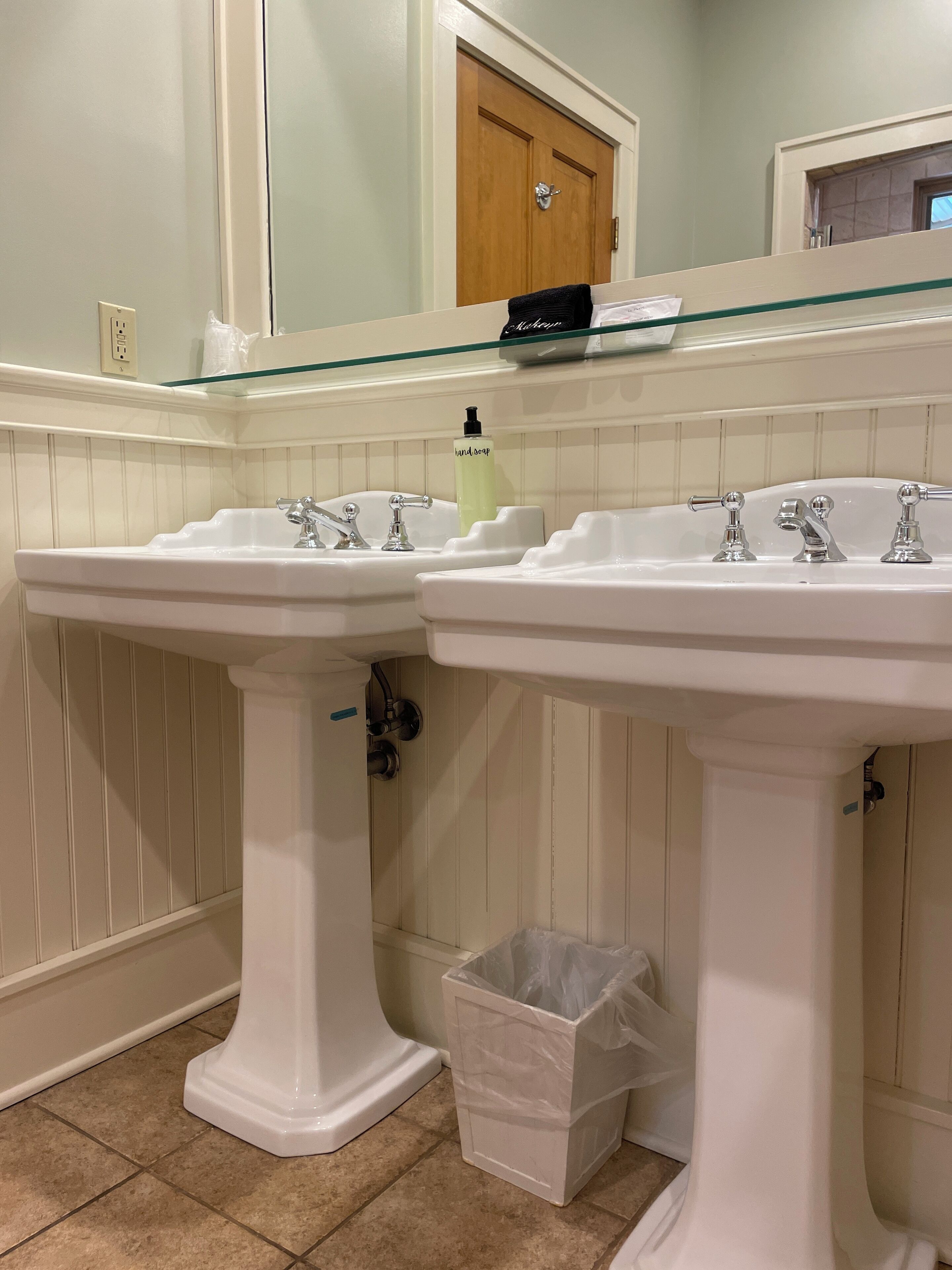 A bathroom with two white pedestal sinks, a glass shelf above, a wooden door, and a trash bin.