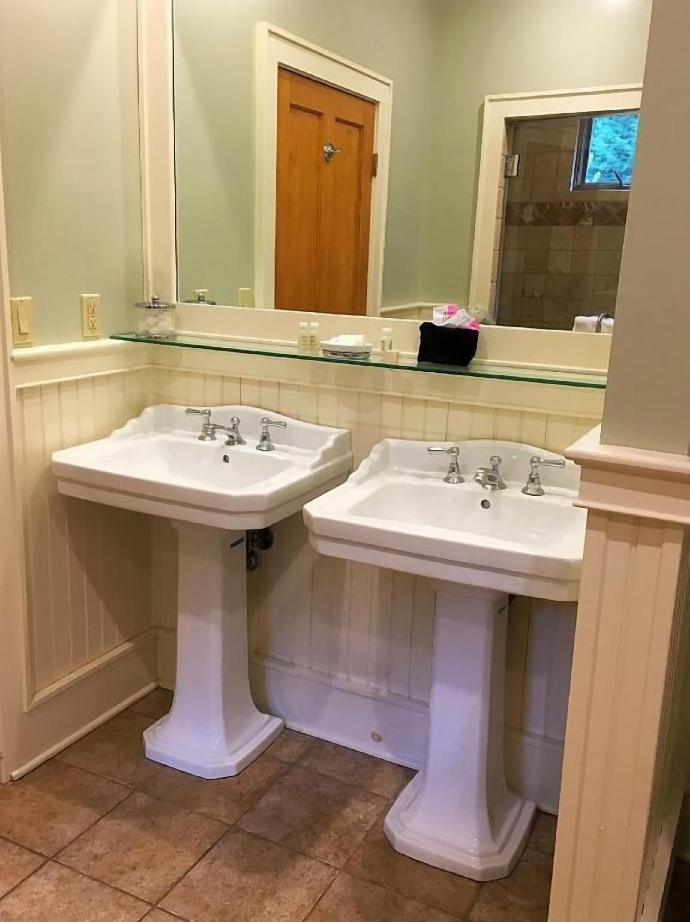 Une salle de bain avec deux lavabos blancs sur pied, une étagère en verre au-dessus des lavabos, une porte en bois et une fenêtre.