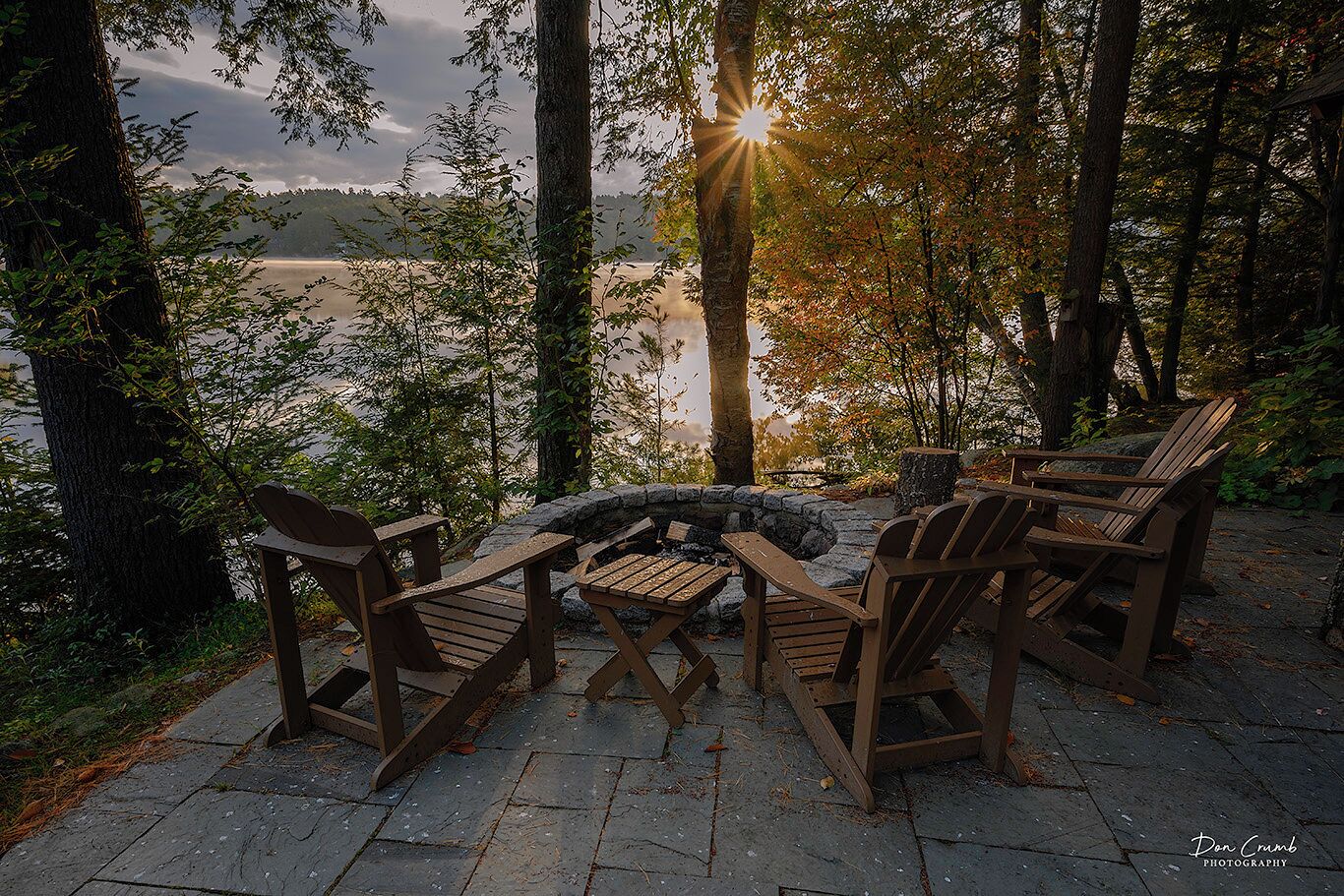 Un hogar de piedra rodeado de sillas Adirondack de madera, sobre un patio de piedra con vistas a un lago y un bosque.