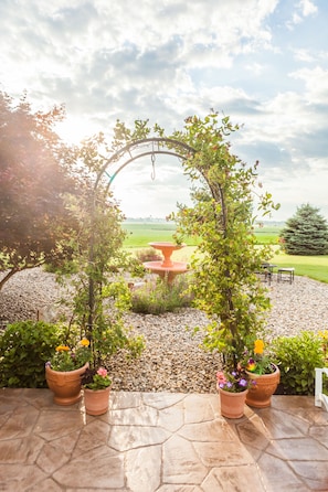 An outdoor patio area with a stone floor, terracotta planters with flowers, a circular table, and a view of a green field.
