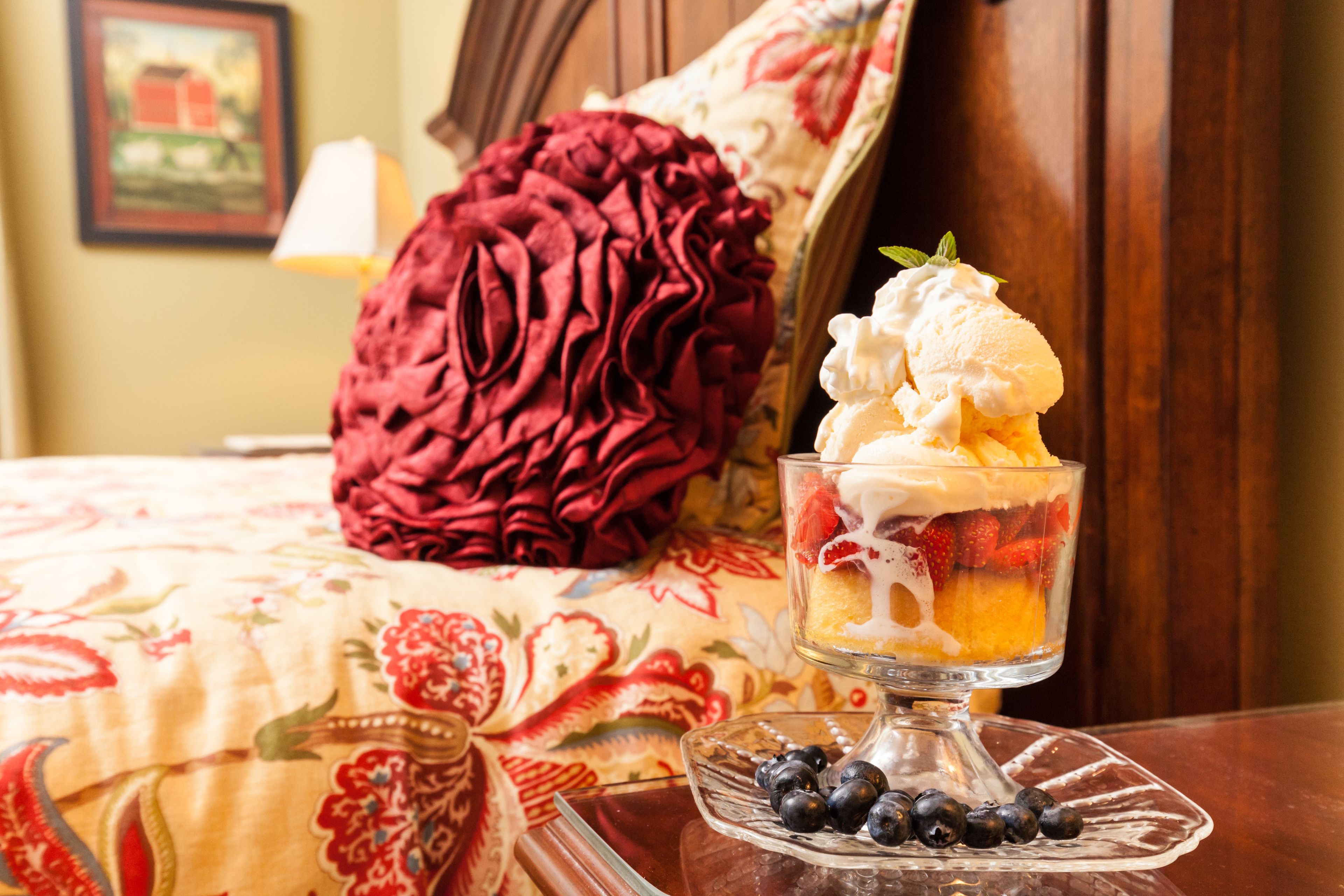 A dessert in a glass bowl on a table, with a bed and a red pillow in the background.