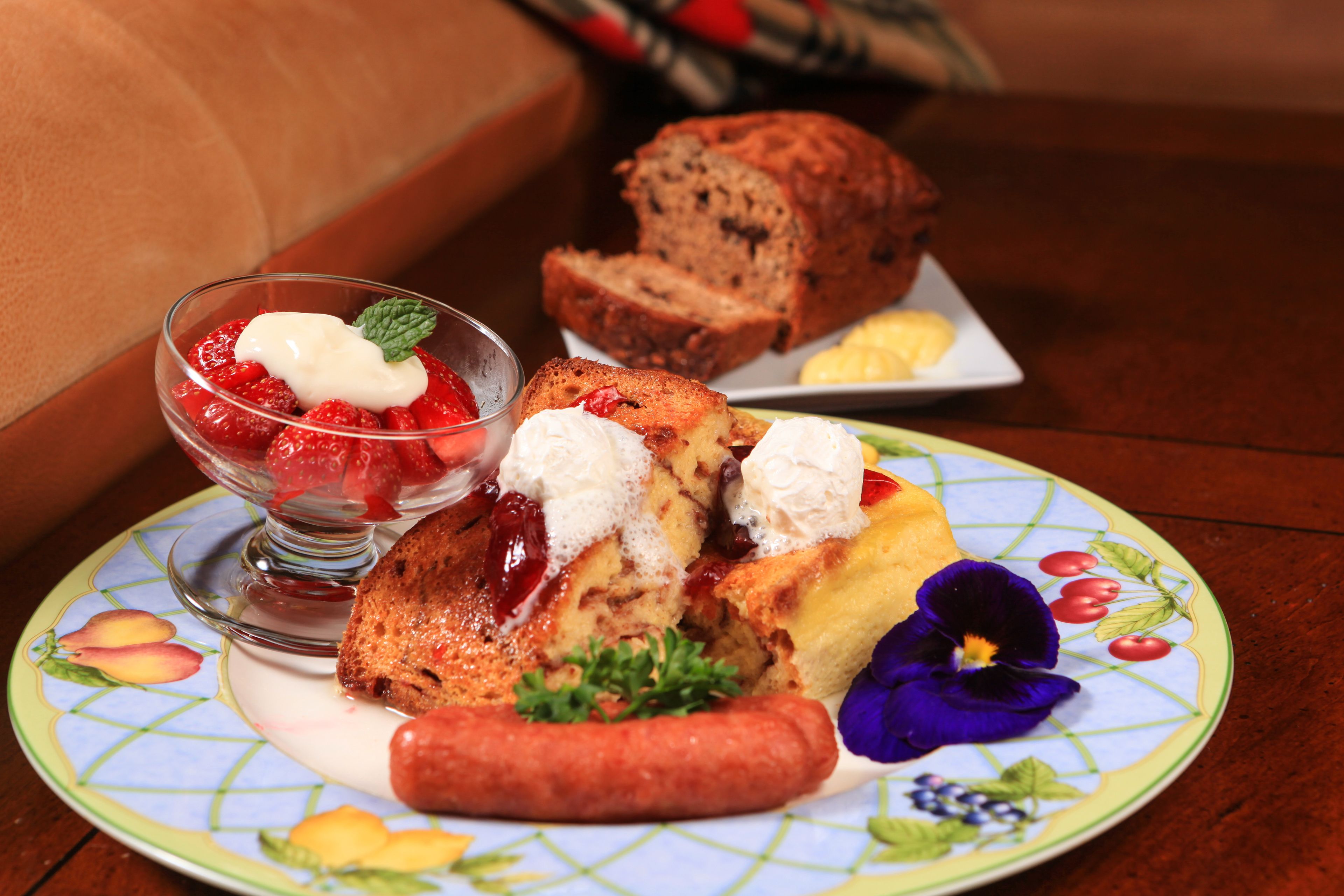 A plate with food items including a sausage, a slice of bread, a piece of cake, and a bowl of strawberries with whipped cream.