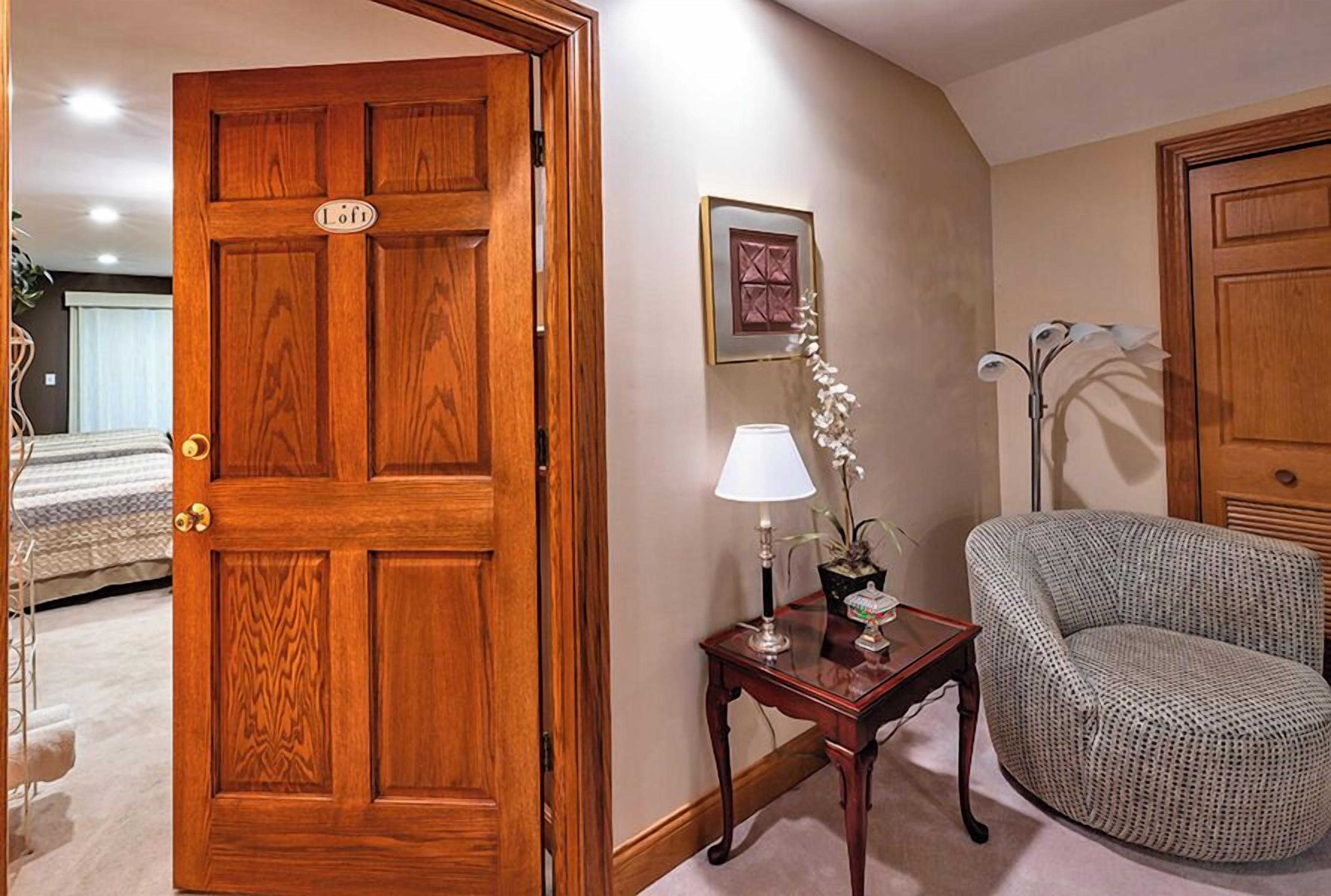 A room with a wooden door labeled "Loft," a small table with a lamp and a plant, a patterned armchair, and a framed picture on the wall.