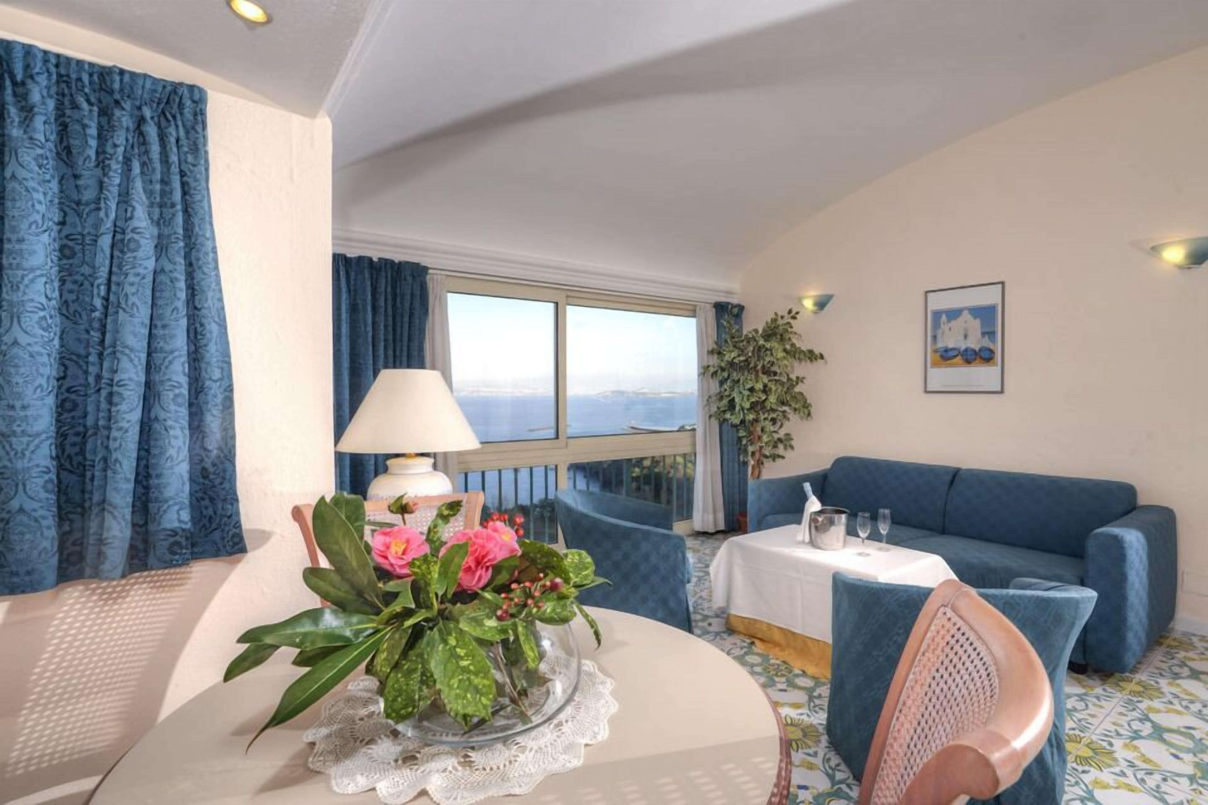 Een kamer met een ronde tafel, een blauwe bank, een raam met uitzicht en een lamp.