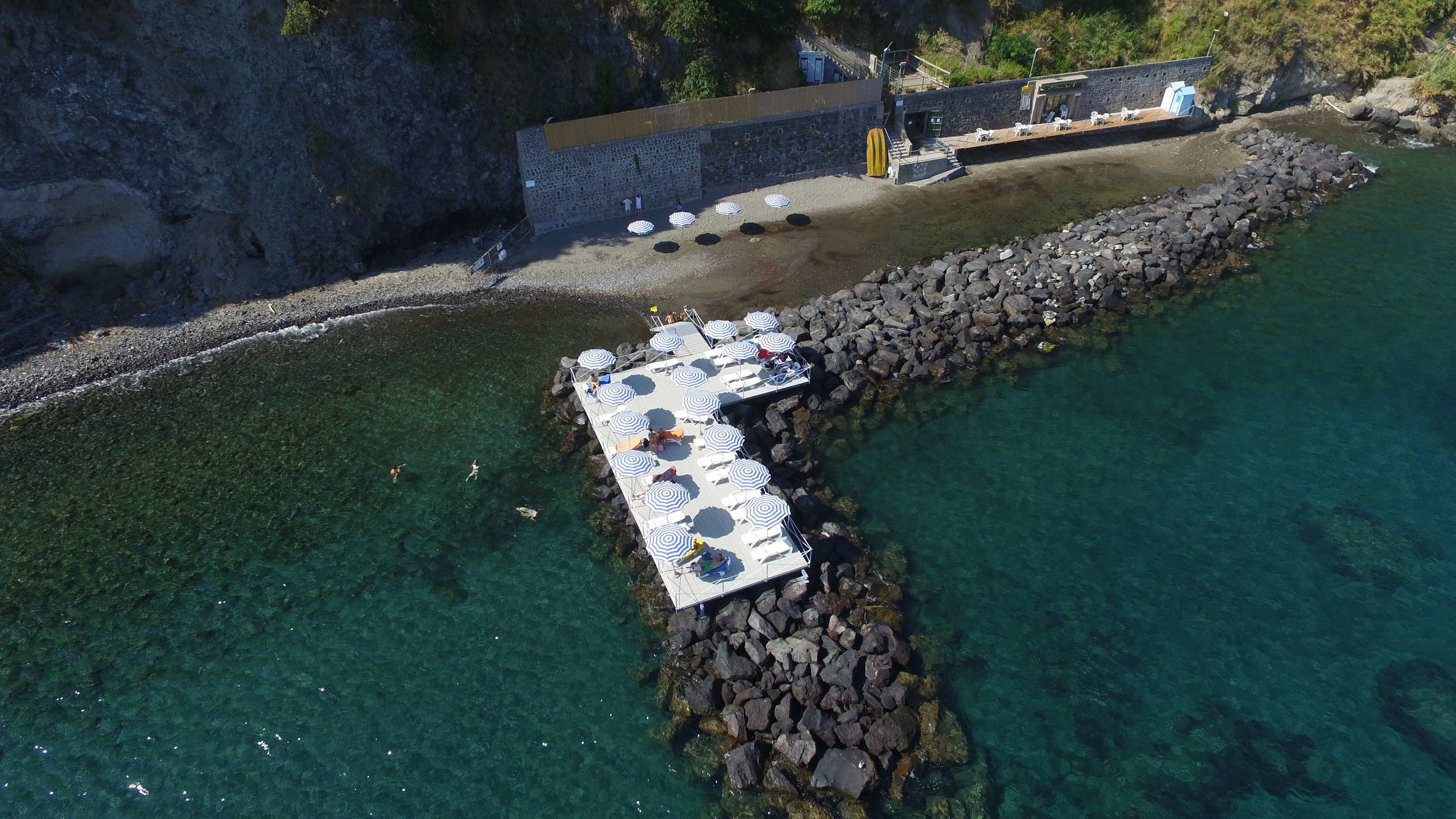 An aerial view of a pier with sun loungers and umbrellas, surrounded by clear water and rocky terrain.