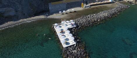 An aerial view of a pier with sun loungers and umbrellas, surrounded by clear water and rocky terrain.