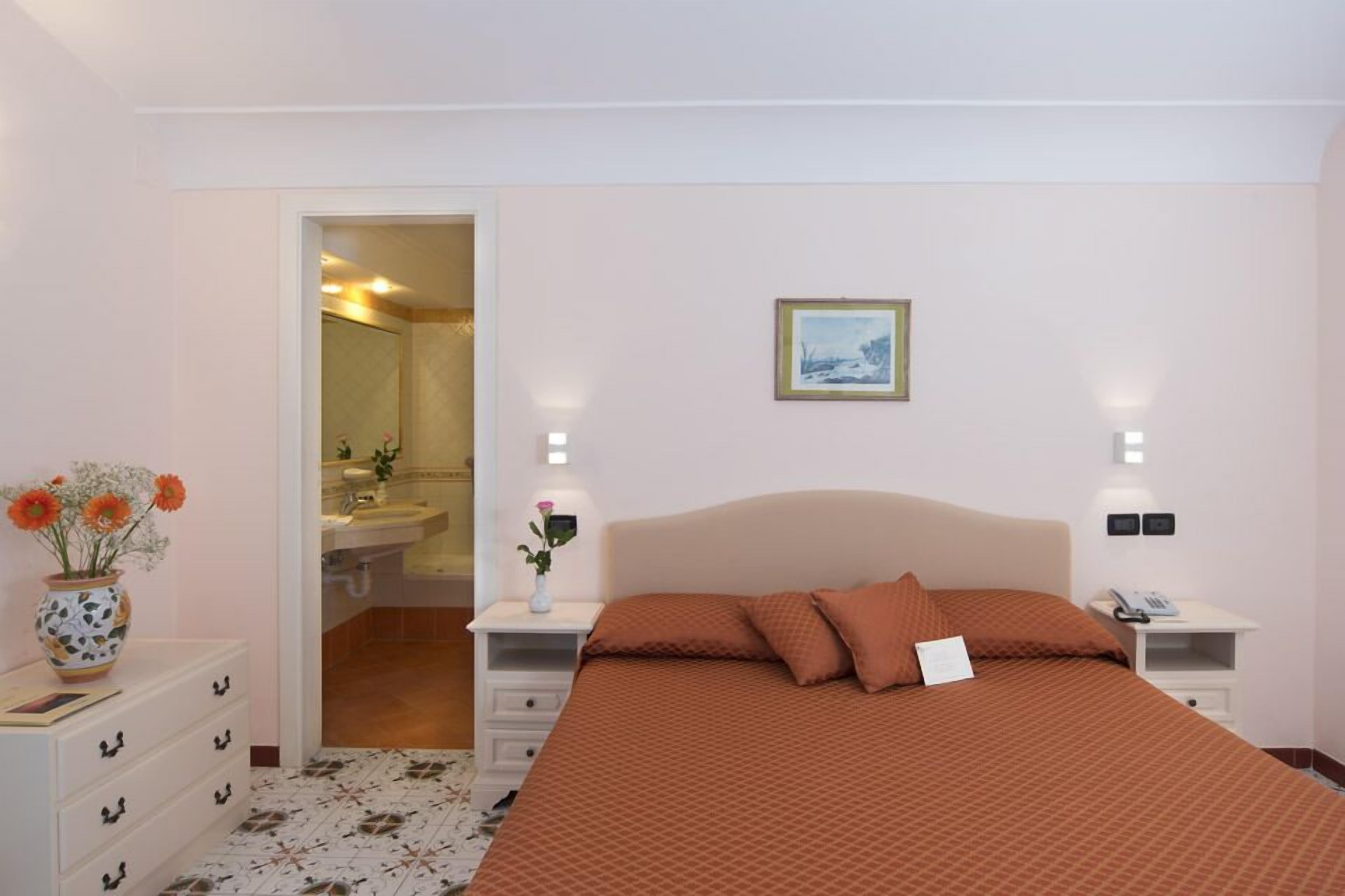 Ett hotellrum med en säng, sängbord, en vas med blommor, en inramad tavla på väggen och ett badrum som syns genom en öppen dörr.