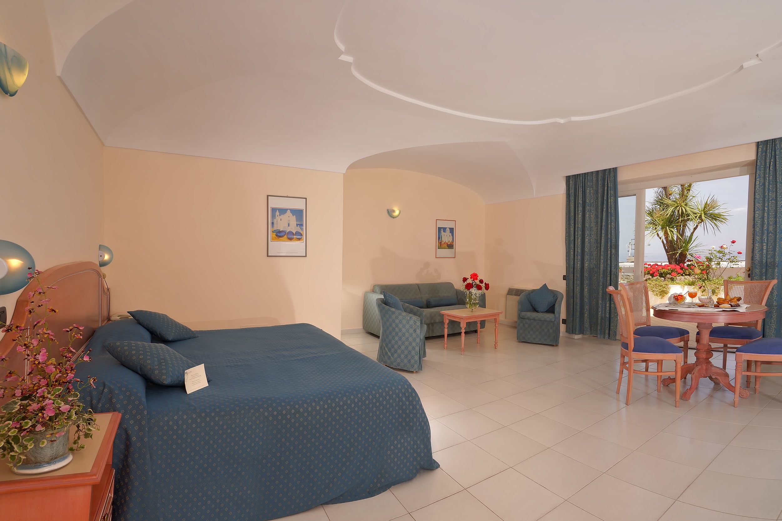 A hotel room with a bed, a sofa, a dining table with chairs, and a large window with curtains.