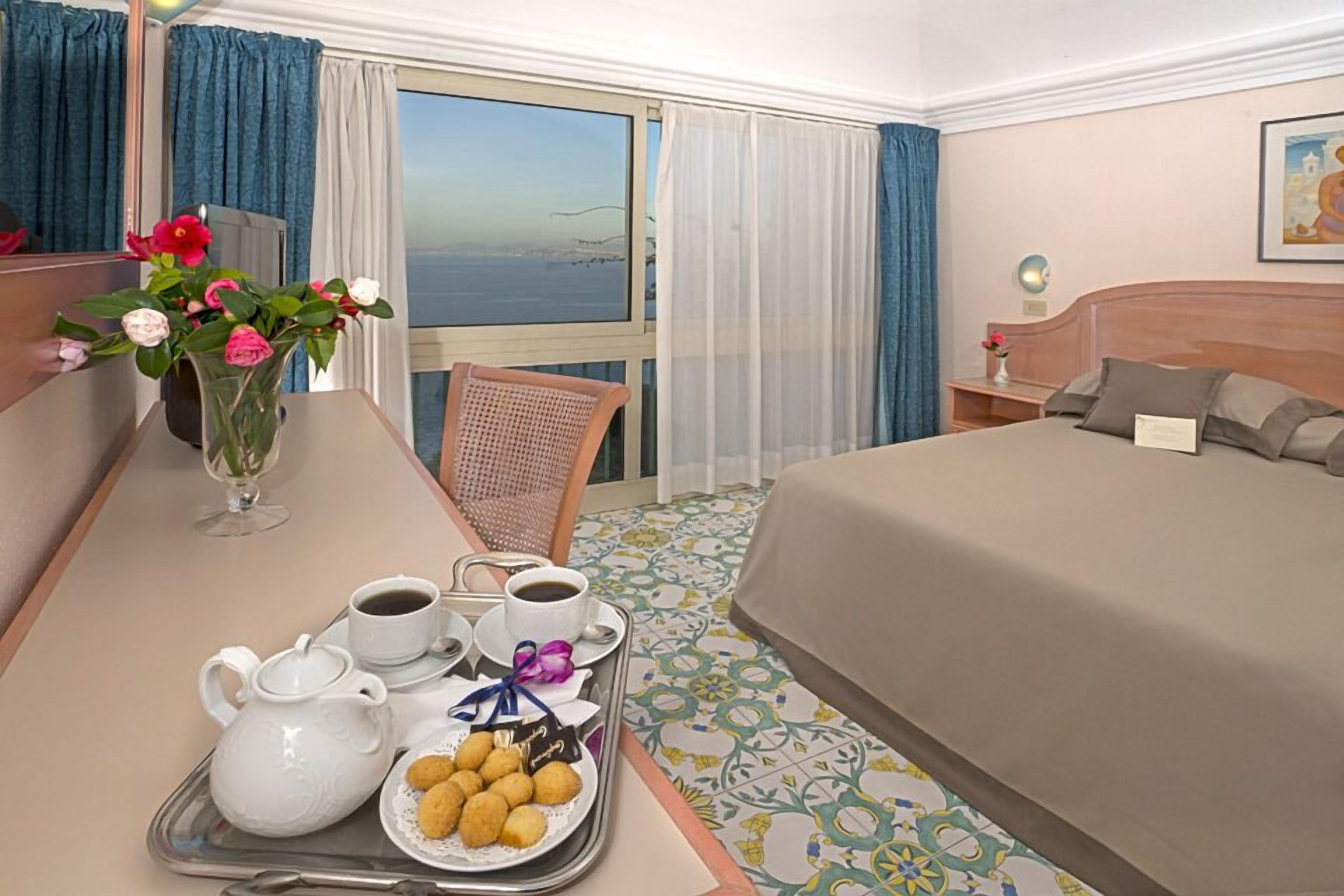 Een hotelkamer met een bed, een eettafel met ontbijtartikelen, een stoel en uitzicht op zee door het raam.