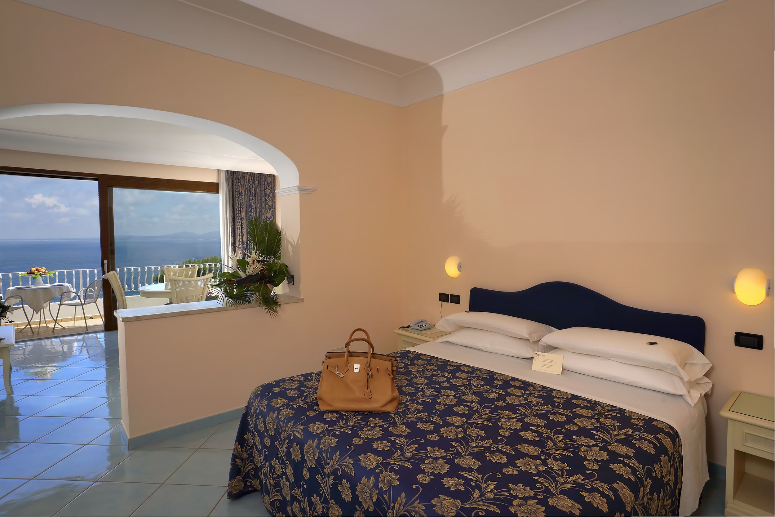 A hotel room with a bed, a nightstand, a window with a view of the sea, and a balcony with outdoor furniture.