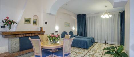 Una habitación con una mesa redonda, dos sillas, una cama con colcha azul, una planta en maceta y un estante en la pared con un jarrón de flores.