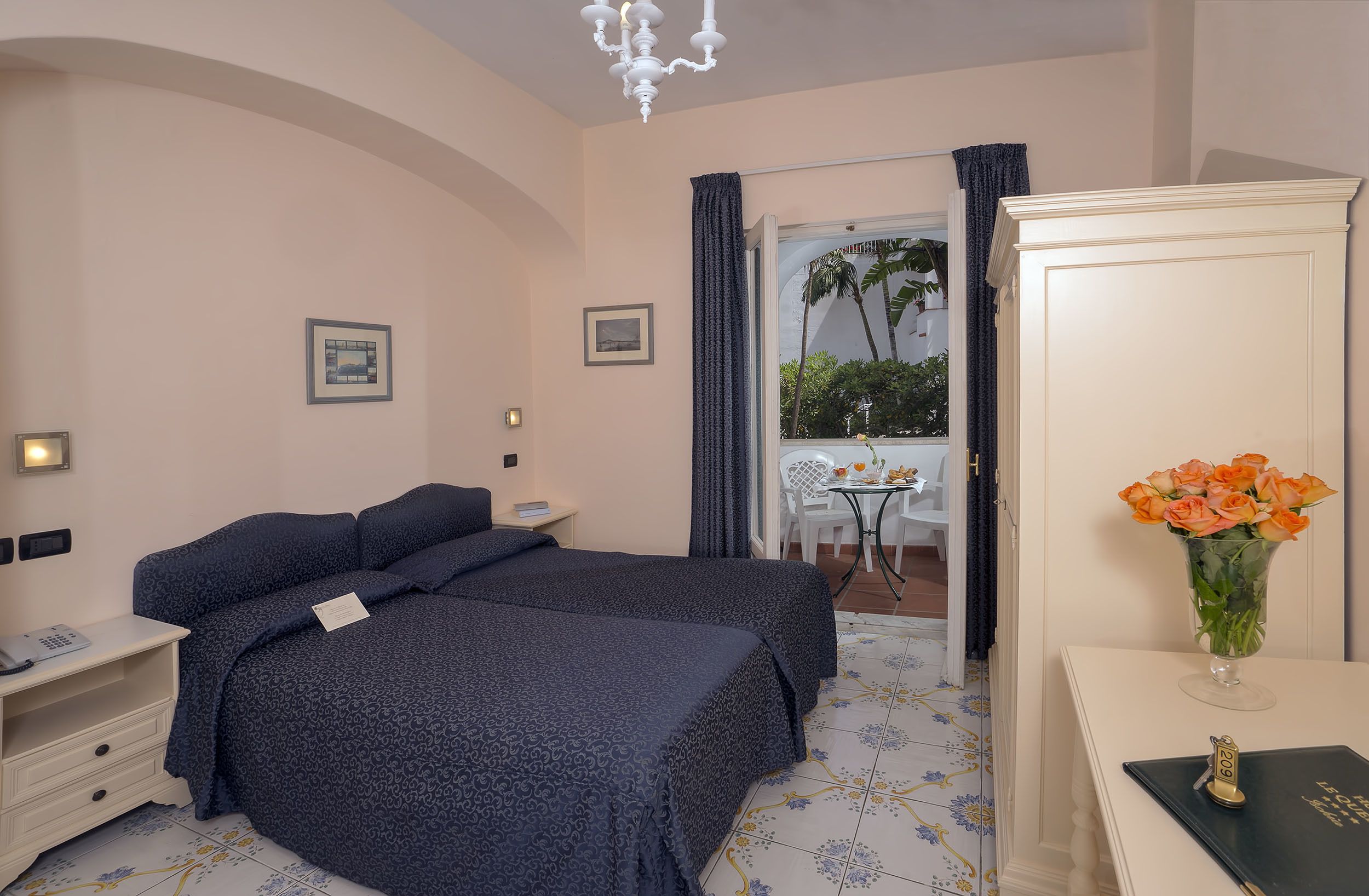 Une chambre d’hôtel avec deux lits, un bureau agrémenté d’un vase de fleurs, une chaise et une vue sur les palmiers par la fenêtre.