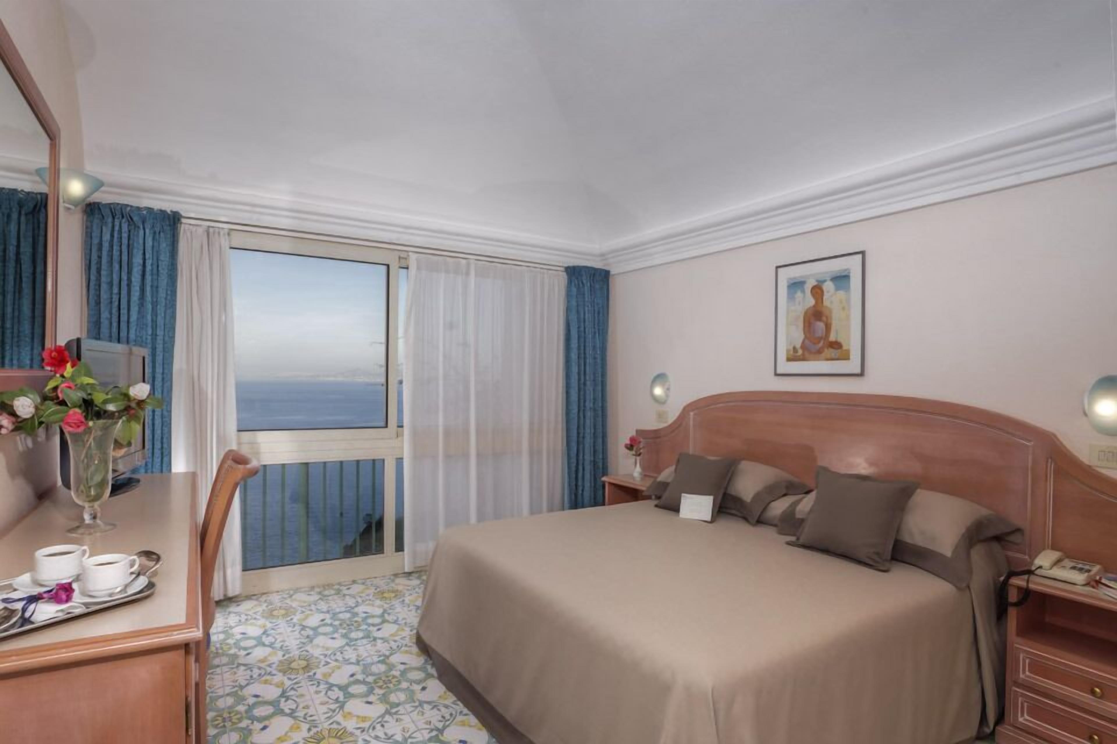 A hotel room with a bed, a desk with a vase of flowers, a view of the sea through a sliding door, and a painting on the wall.