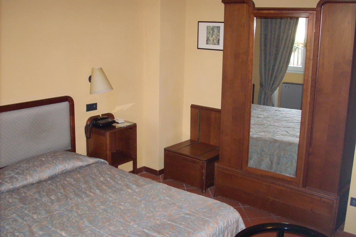 A hotel room with a bed, a bedside table with a lamp, a small desk, and a mirror.