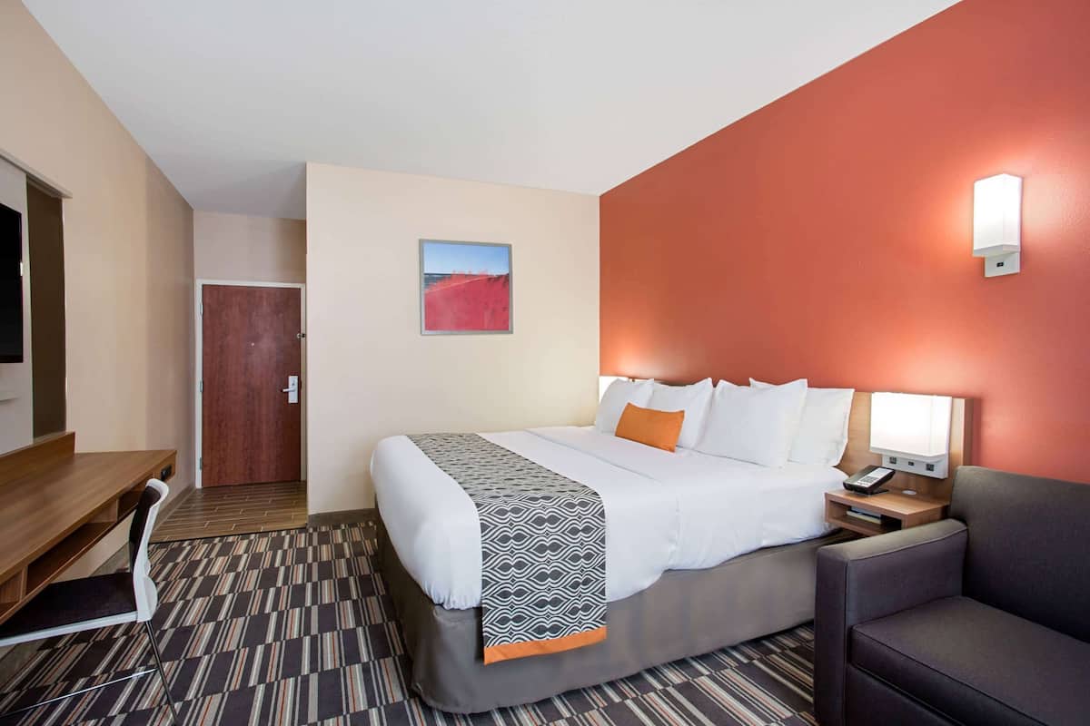 A hotel room with a bed, a desk, a chair, a nightstand, and a wall-mounted lamp.
