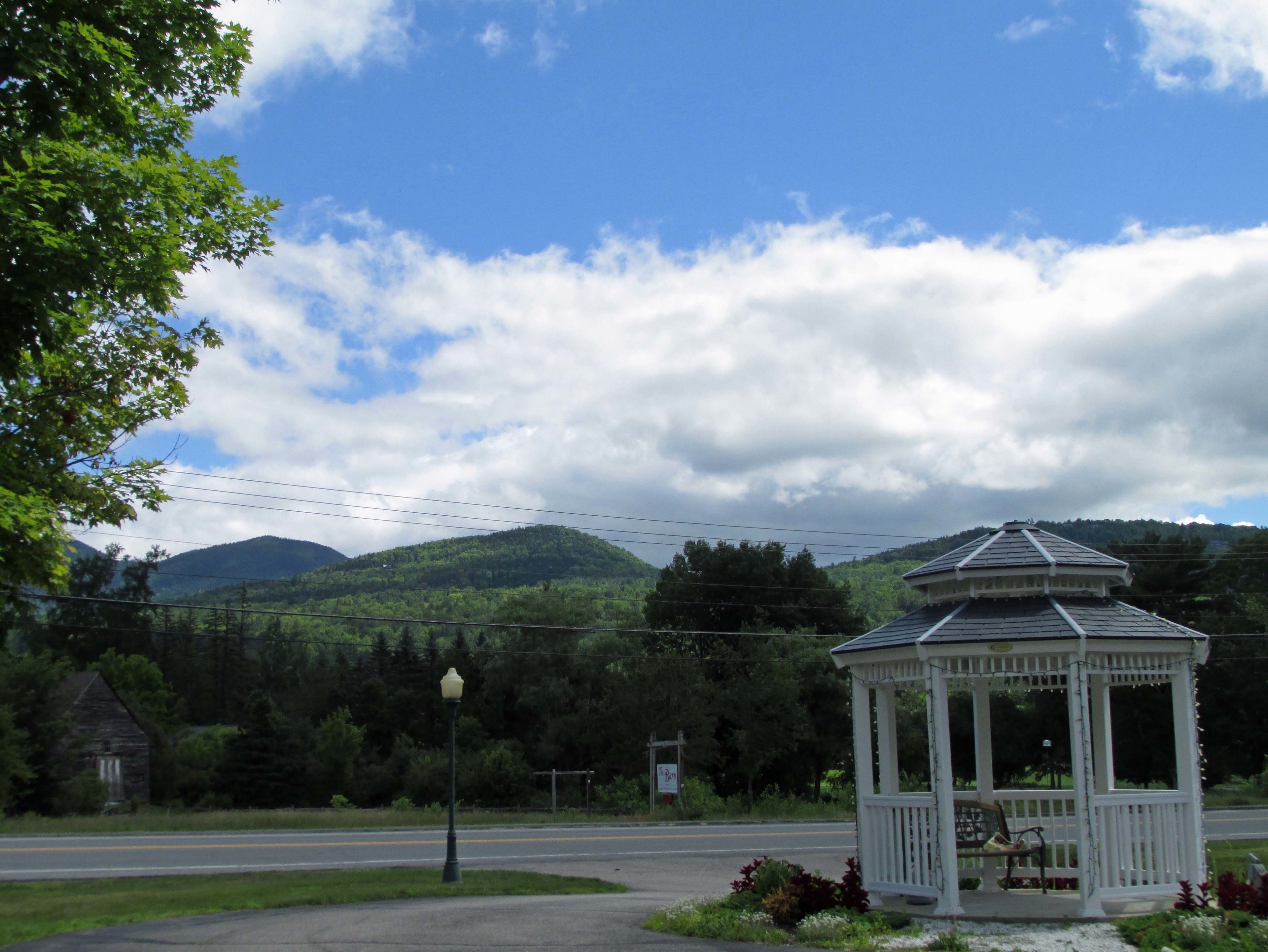 A gazebo with a white exterior and a small porch, located beside a road with a street lamp and a sign in the background.