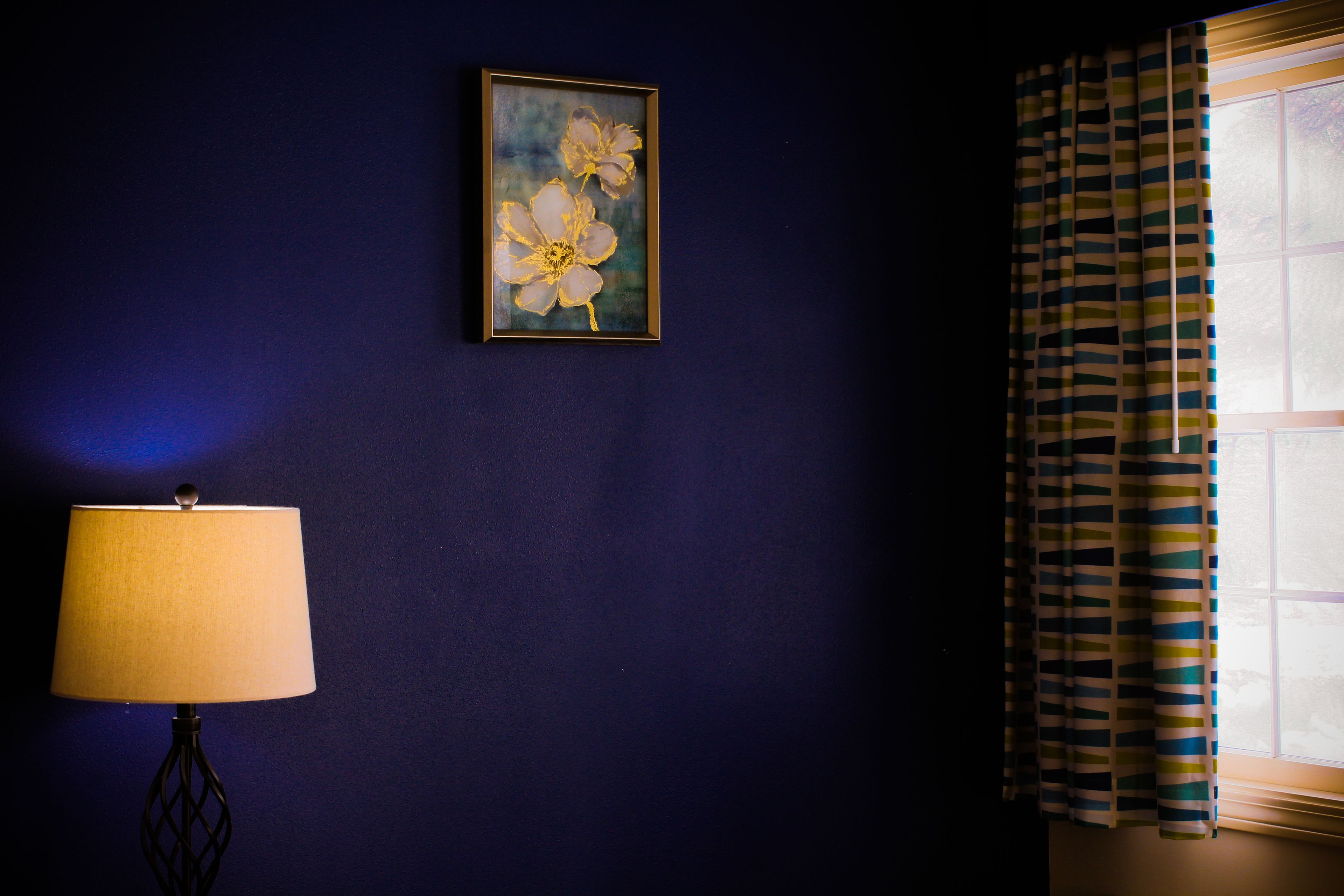A room with a dark blue wall, a framed painting of flowers, a lamp, and a window with striped curtains.