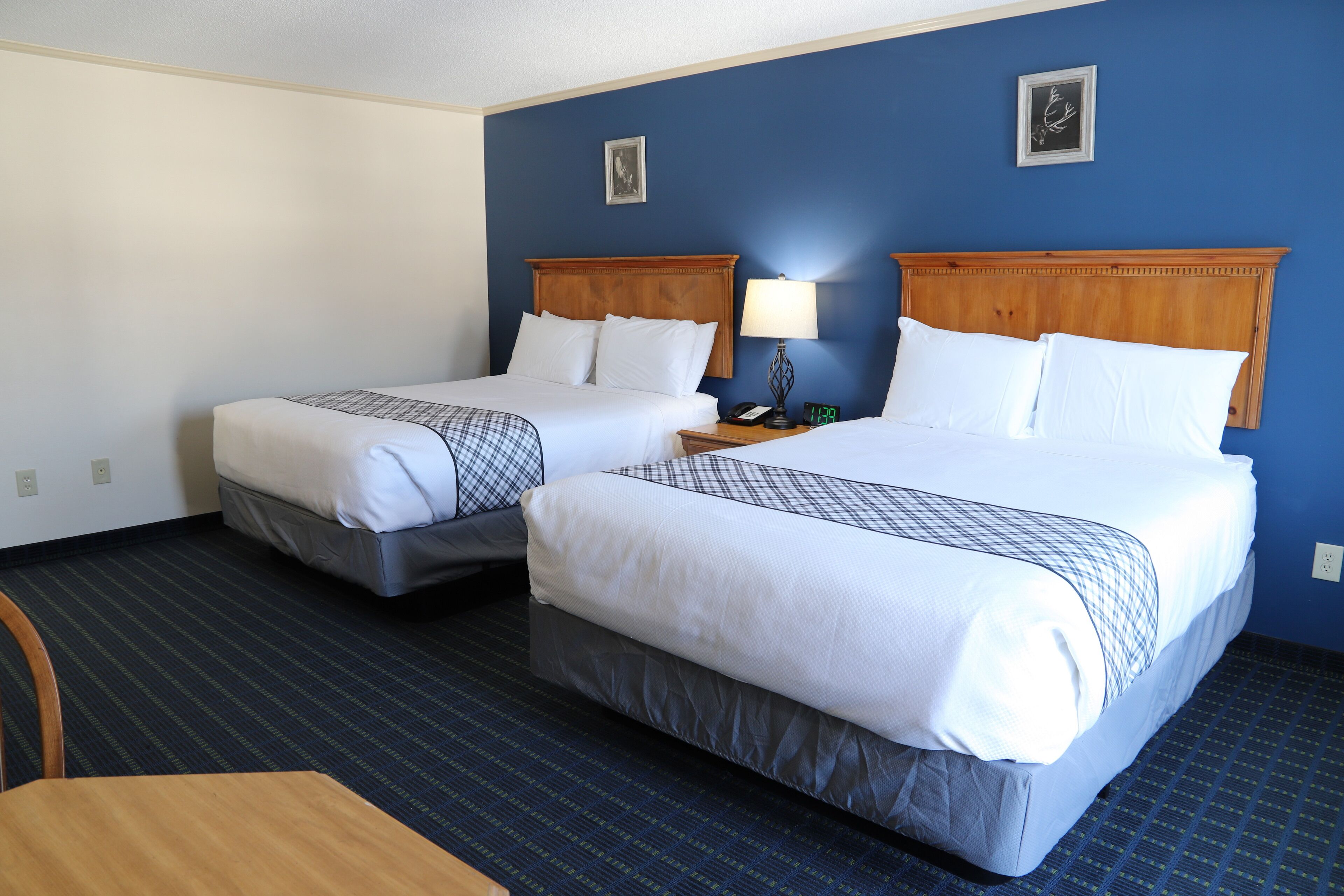 Habitación de hotel con dos camas, cabecero de madera, mesita de noche con lámpara y una pequeña mesa.