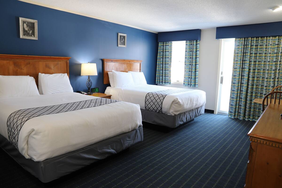 A hotel room with two beds, a wooden headboard, a nightstand with a lamp, and a desk with a chair.