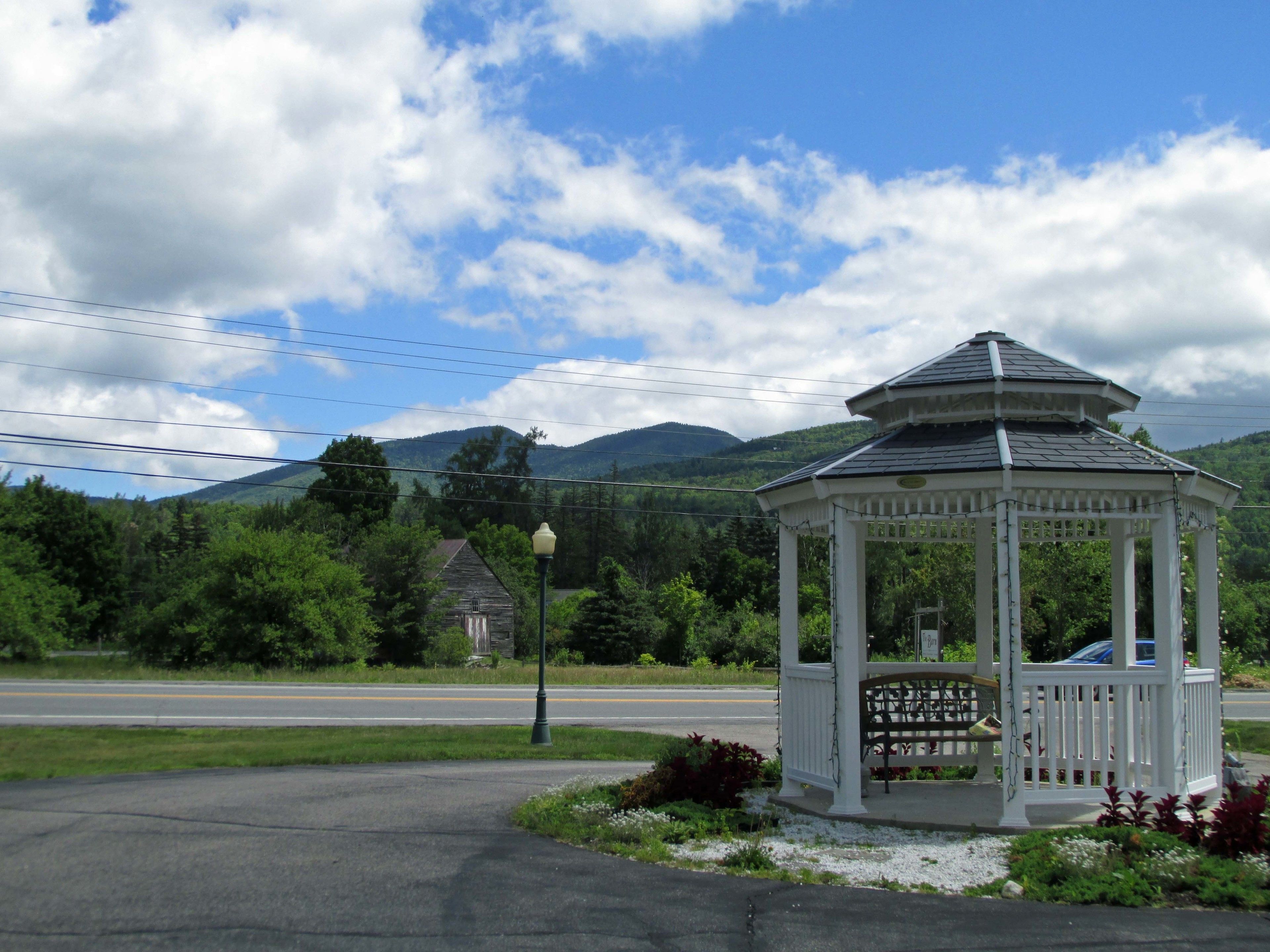 A gazebo with a white structure and a roof, surrounded by greenery and a road.