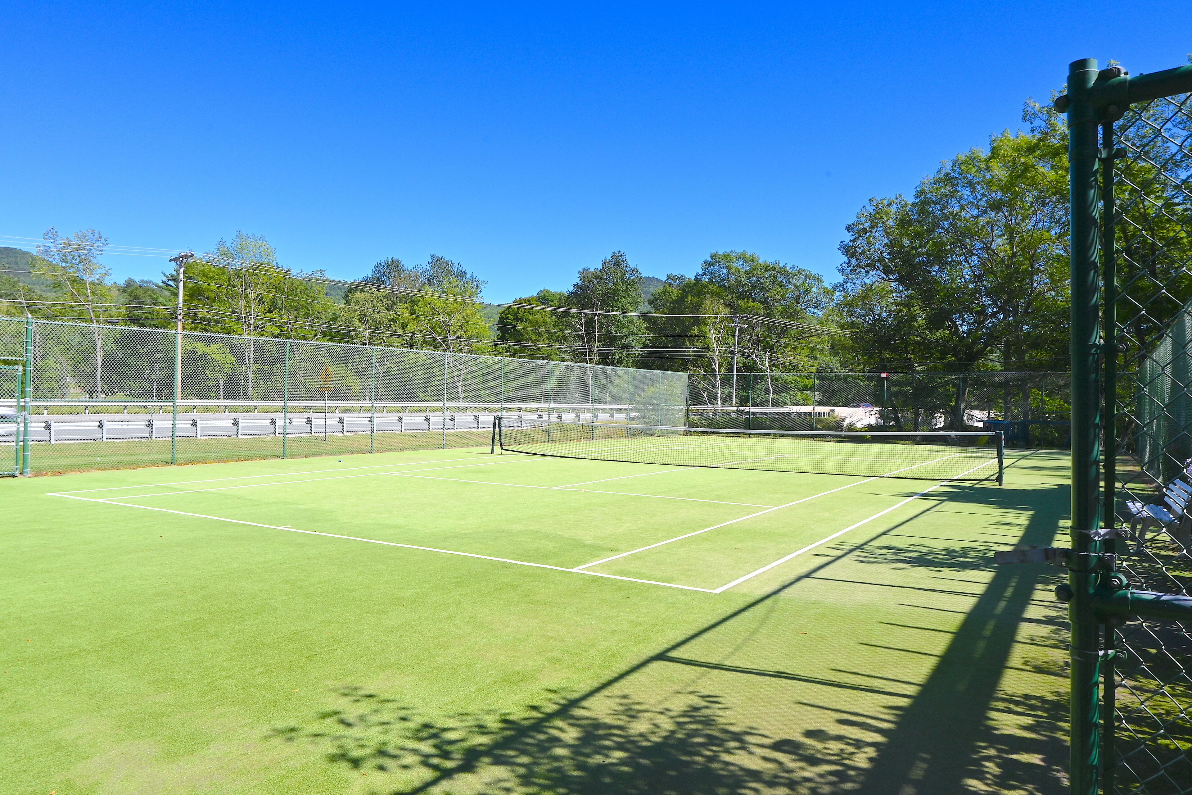 Una cancha de tenis con red, rodeada por una cerca de malla y árboles al fondo.