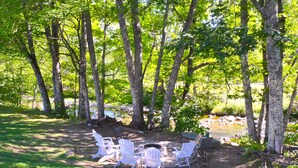 A campsite with white Adirondack chairs arranged around a fire pit, surrounded by trees and a small stream. - The Lodge At Jackson Village (Jackson)