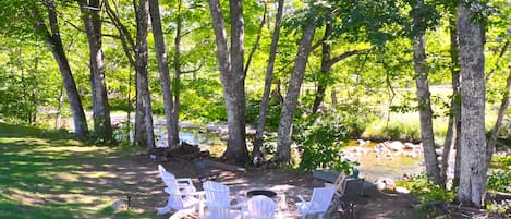 A campsite with white Adirondack chairs arranged around a fire pit, surrounded by trees and a small stream.