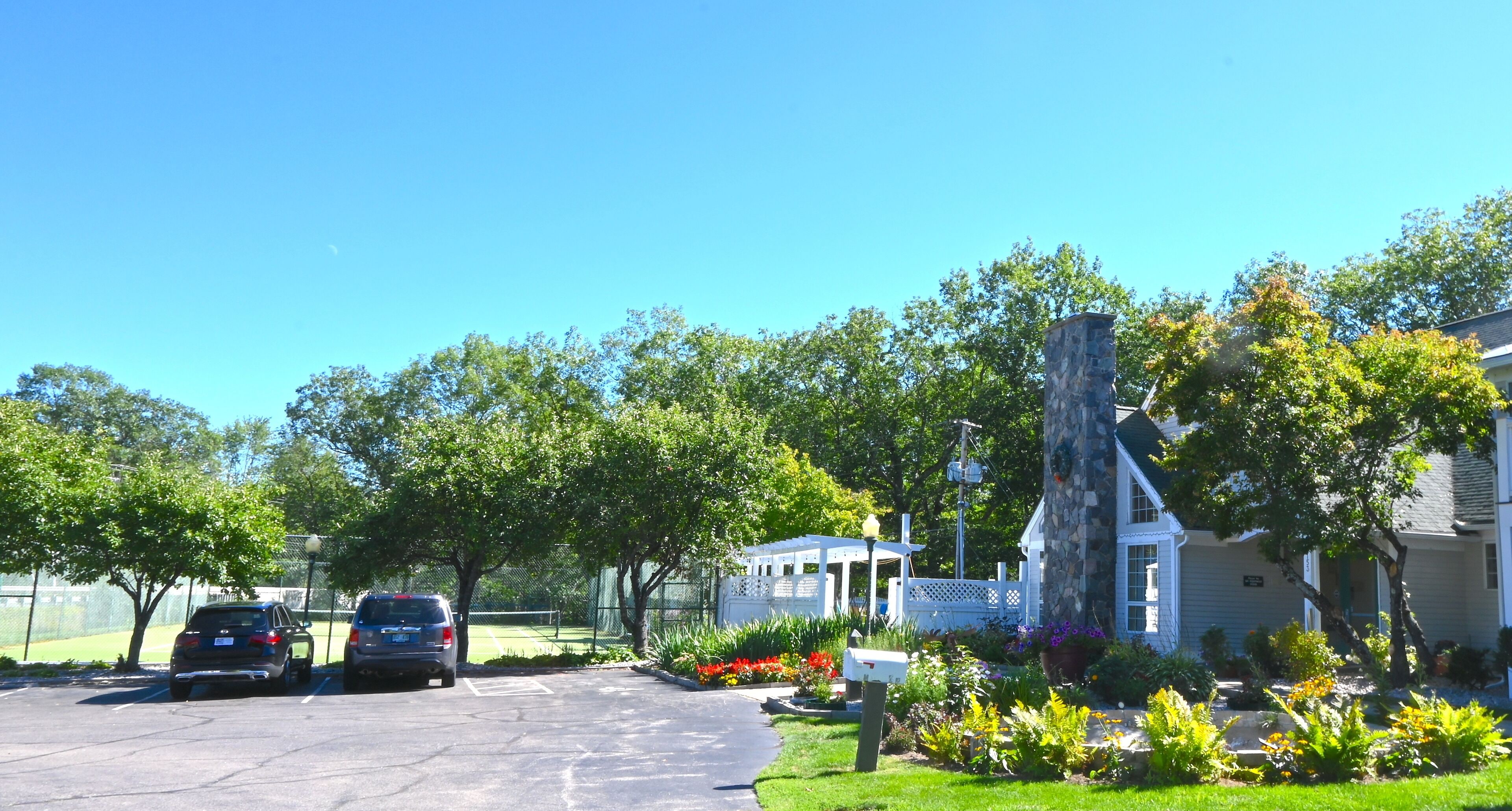 A parking lot with two cars, a gazebo, a stone pillar, and a house with a white fence.