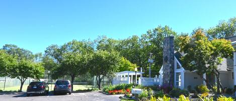 A parking lot with two cars, a gazebo, a stone pillar, and a house with a white fence.