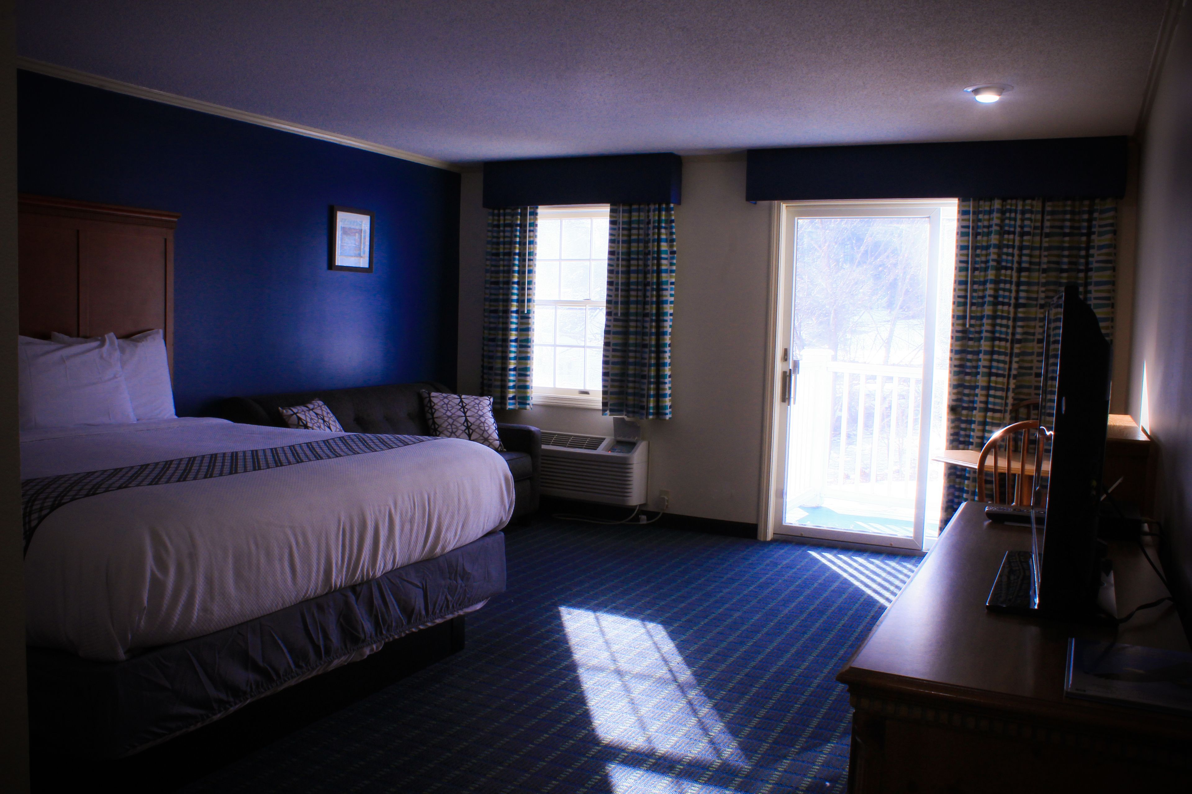 A hotel room with a large bed, a desk with a computer, a window with curtains, and a door leading to a balcony.