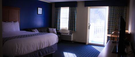 Habitación de hotel con una cama grande, un escritorio con computadora, una ventana con cortinas y una puerta que da a un balcón.