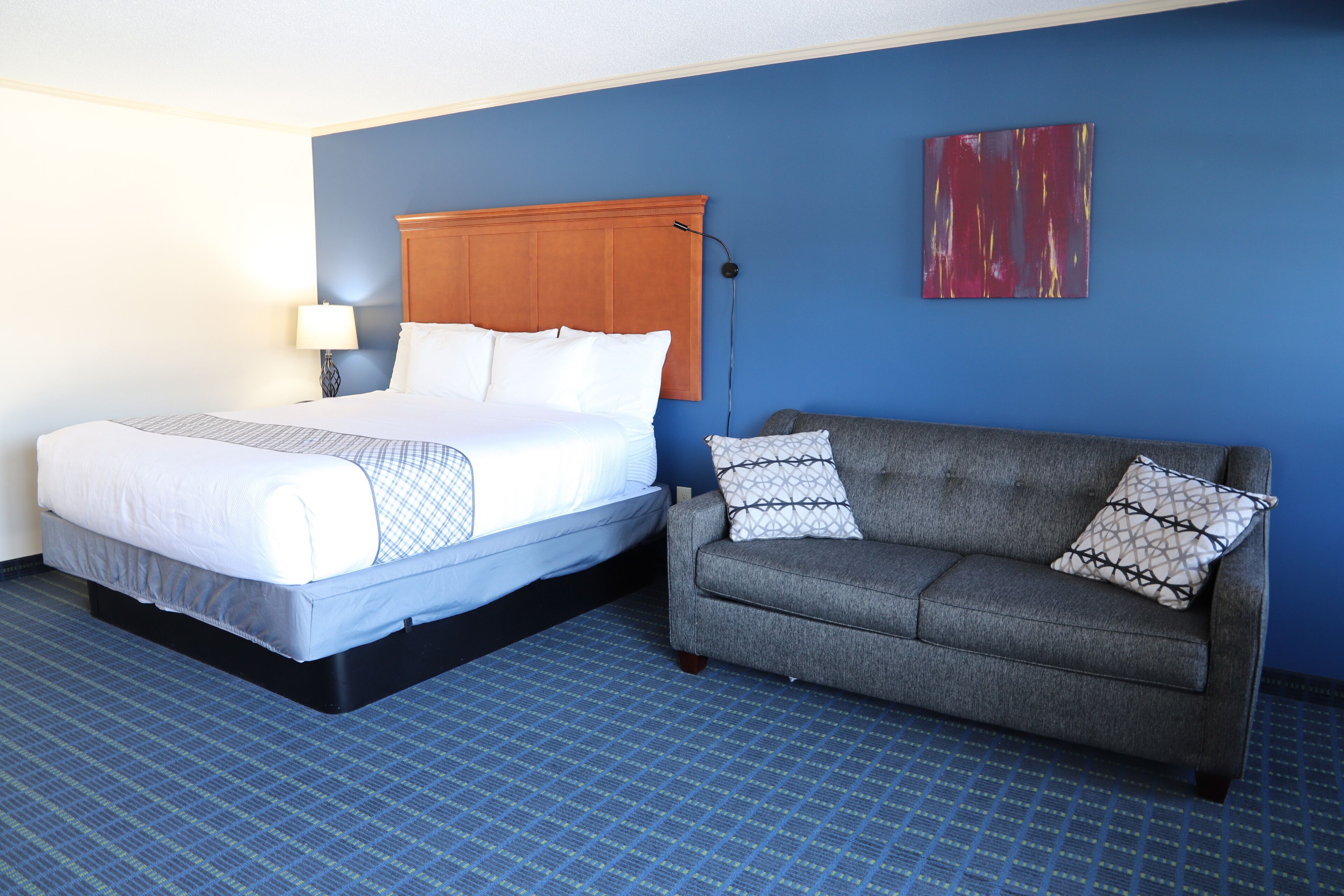 A hotel room with a bed, a grey sofa, a blue wall, and a red abstract painting.