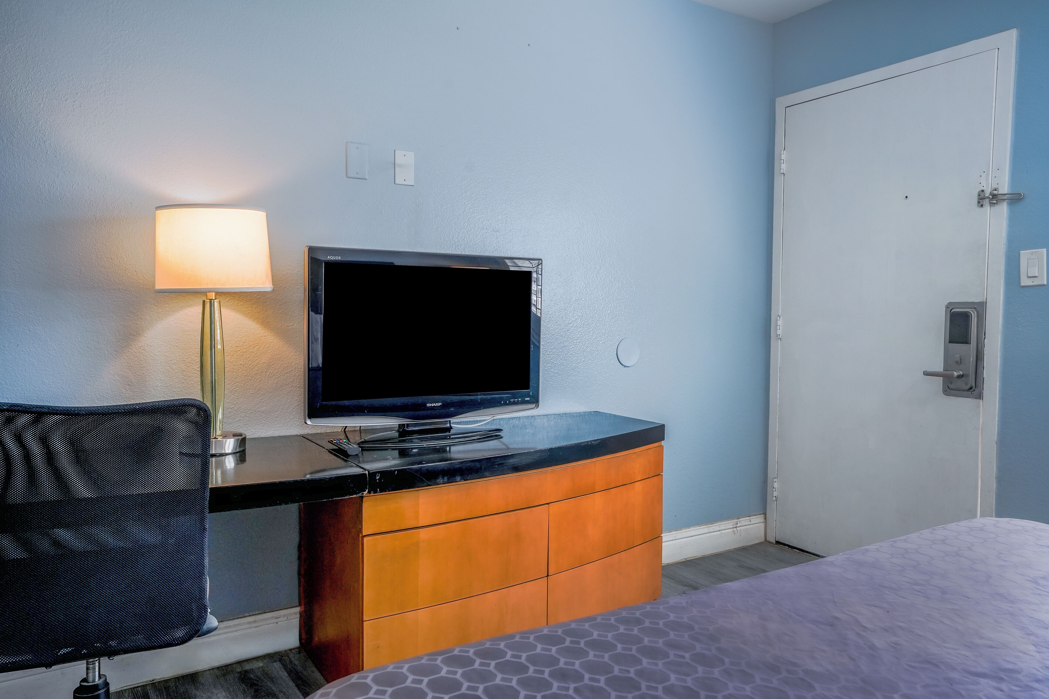 A hotel room with a bed, a desk with a television, a chair, and a door.