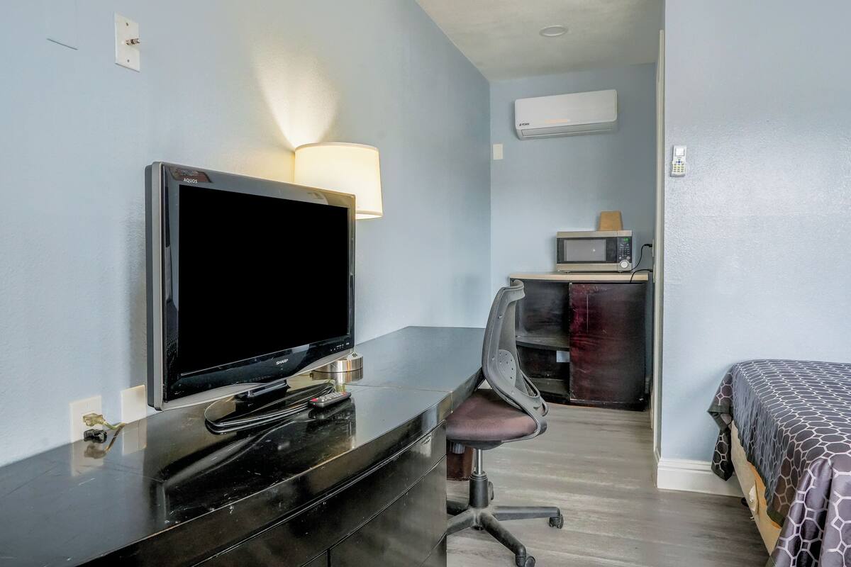 A hotel room with a flat-screen TV on a dresser, a desk chair, a bed with a patterned bedspread, a microwave, and a wall-mounted phone.