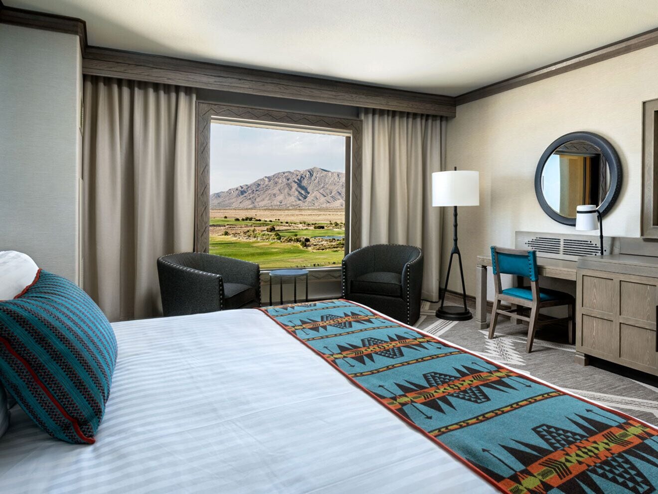 A hotel room with a large bed, a desk, a chair, a lamp, and a view of a mountainous landscape.