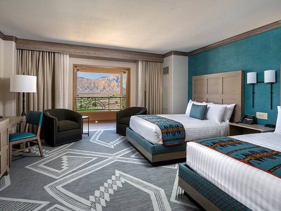 A hotel room with two beds, a desk, a chair, a large window with a mountain view, and a patterned rug.