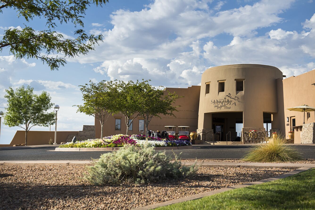 A building with a curved facade, a landscaped entrance area with trees and flowers, and a clear sky.