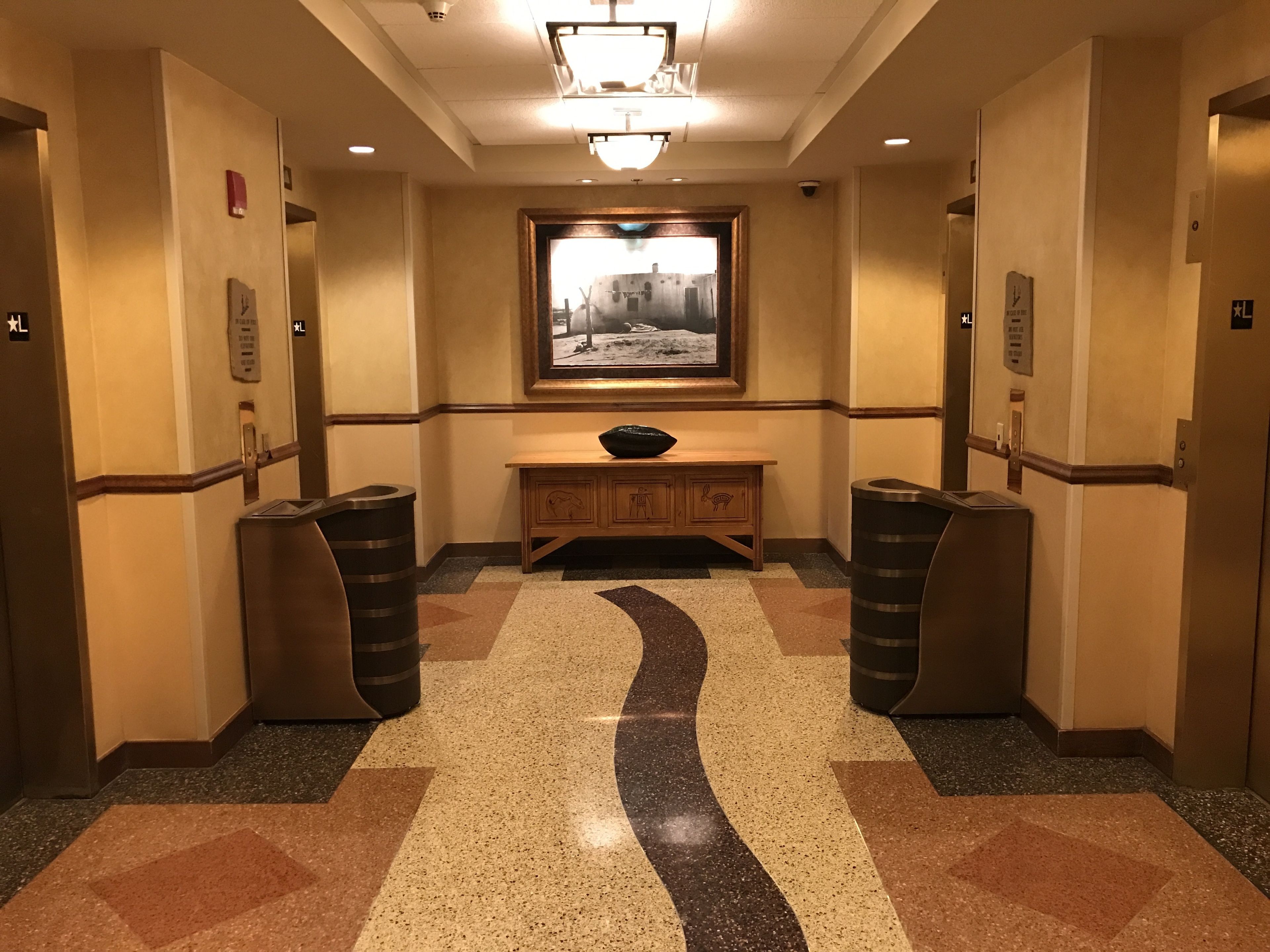 A hotel lobby with a central wooden desk, two trash bins, a framed picture on the wall, and a patterned floor.