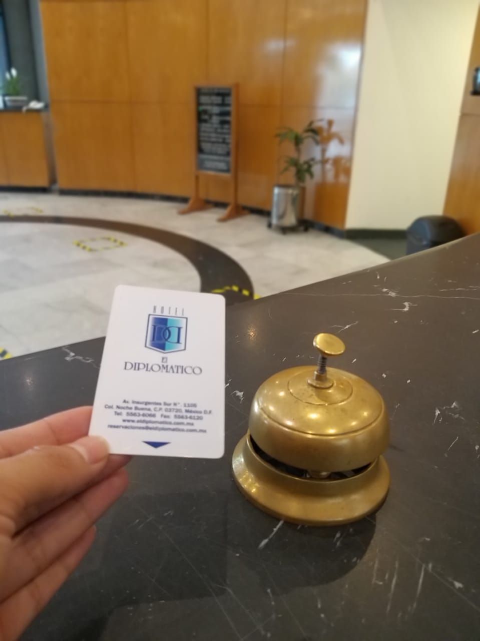 Une main tenant une carte de visite sur laquelle figure le nom « DIPLOMATICO », et une clochette d’hôtel posée sur une table.