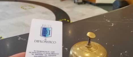 Une main tenant une carte de visite sur laquelle figure le nom « DIPLOMATICO », et une clochette d’hôtel posée sur une table.
