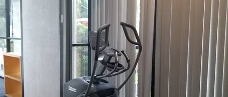 Un vélo d’appartement doté d’un écran numérique, placé près d’une fenêtre munie de stores.