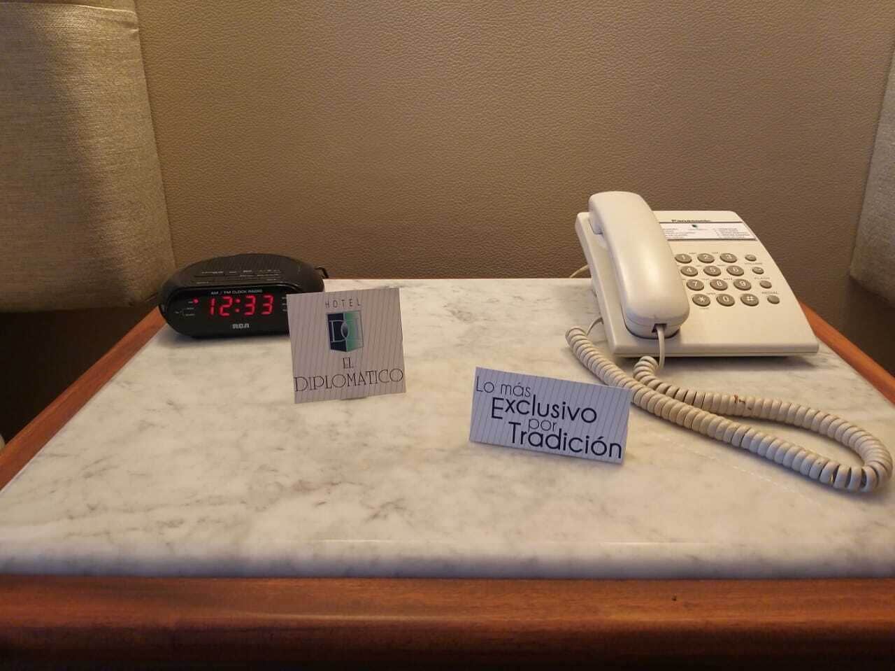 A hotel room with a marble table, a clock, a traditional phone, and a sign indicating 'Hotel Diplomático'.