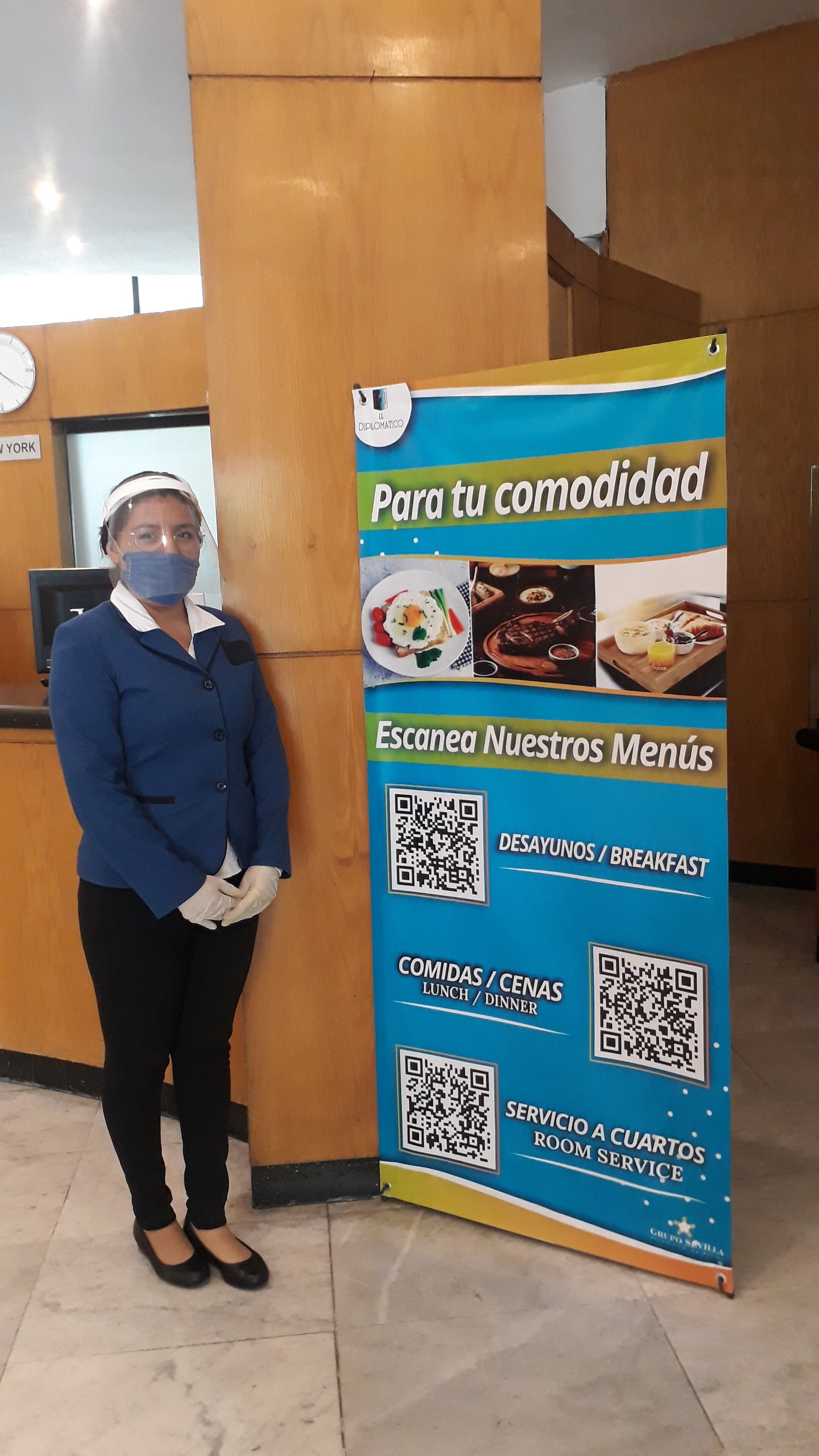 A hotel reception area with a staff member standing next to a menu board displaying QR codes for breakfast, lunch, dinner, and room service.