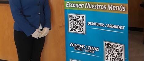 A hotel reception area with a staff member standing next to a menu board displaying QR codes for breakfast, lunch, dinner, and room service.