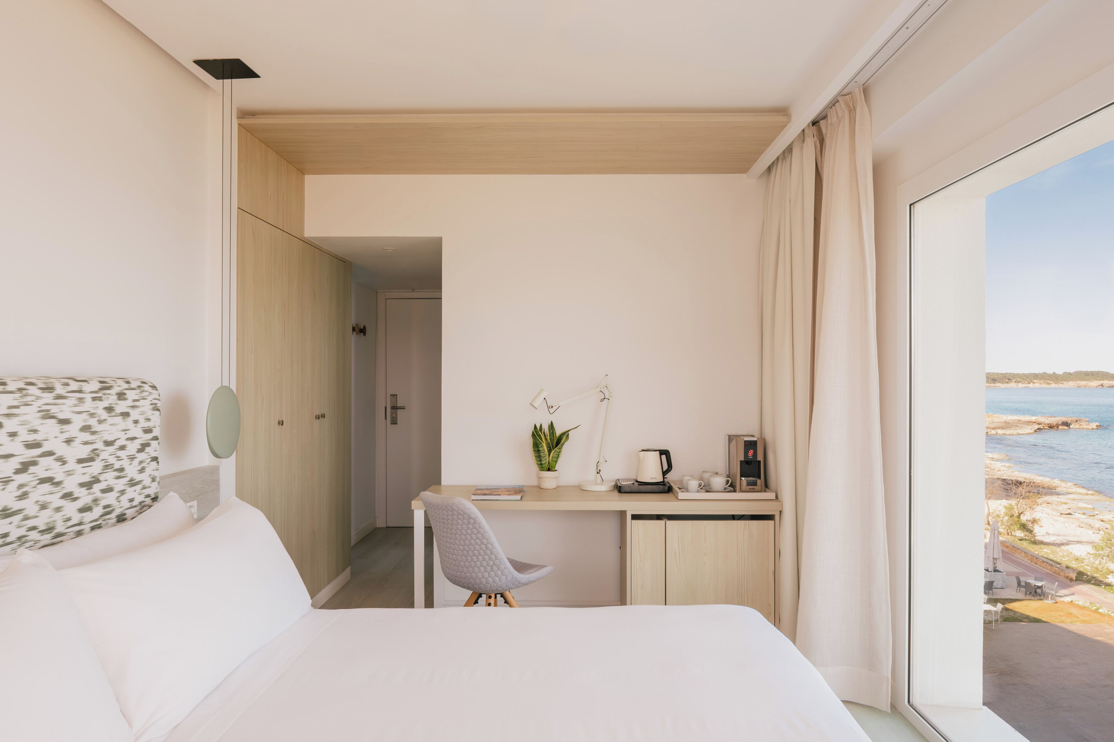 Habitación de hotel con cama, escritorio con silla, planta en maceta, cafetera y vista al mar.