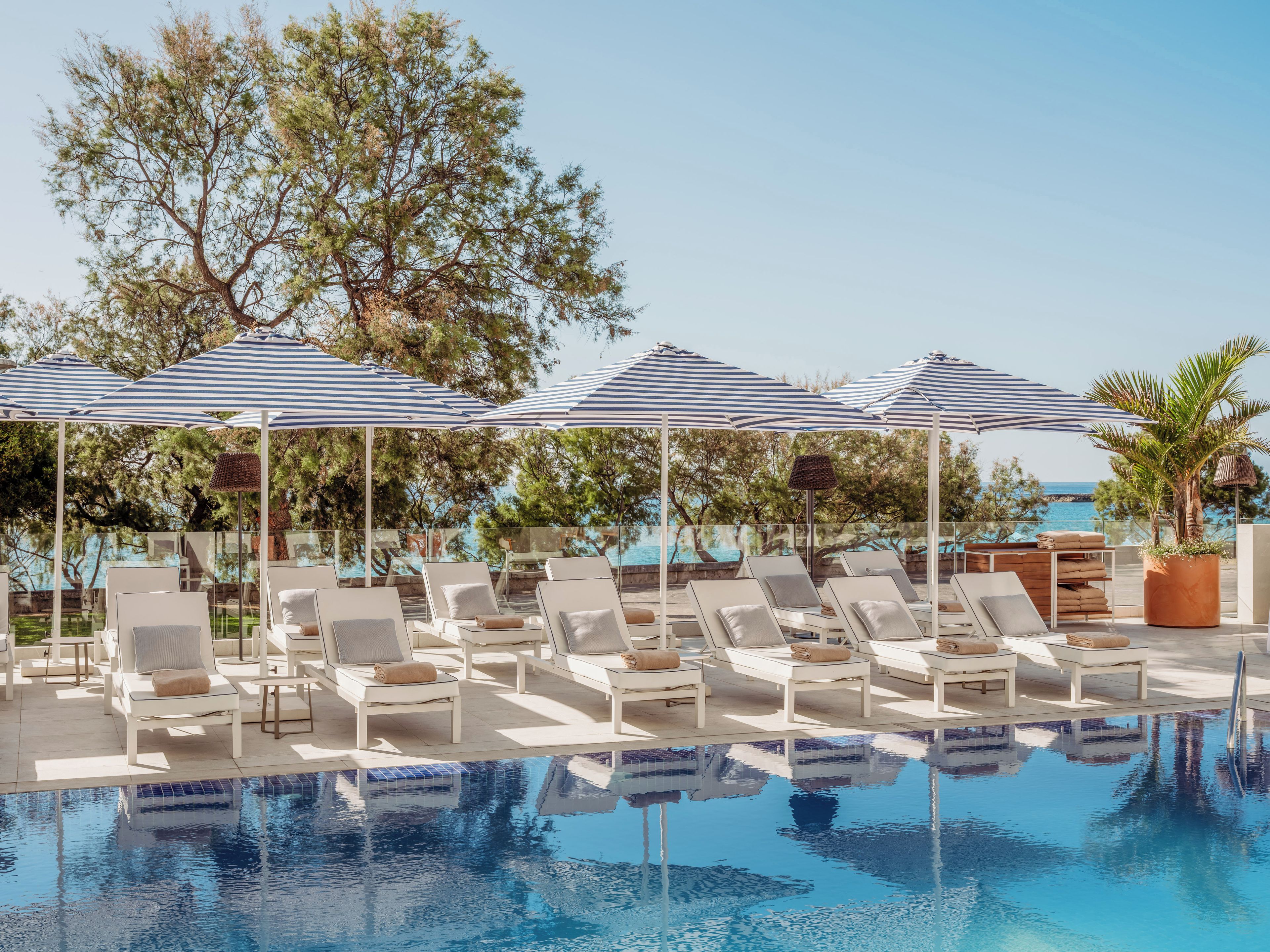 A poolside area with lounge chairs and umbrellas, a clear view of the sea, and a reflection in the pool.