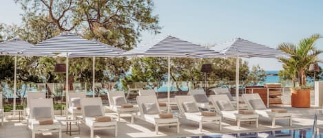A poolside area with lounge chairs and umbrellas, a clear view of the sea, and a reflection in the pool.