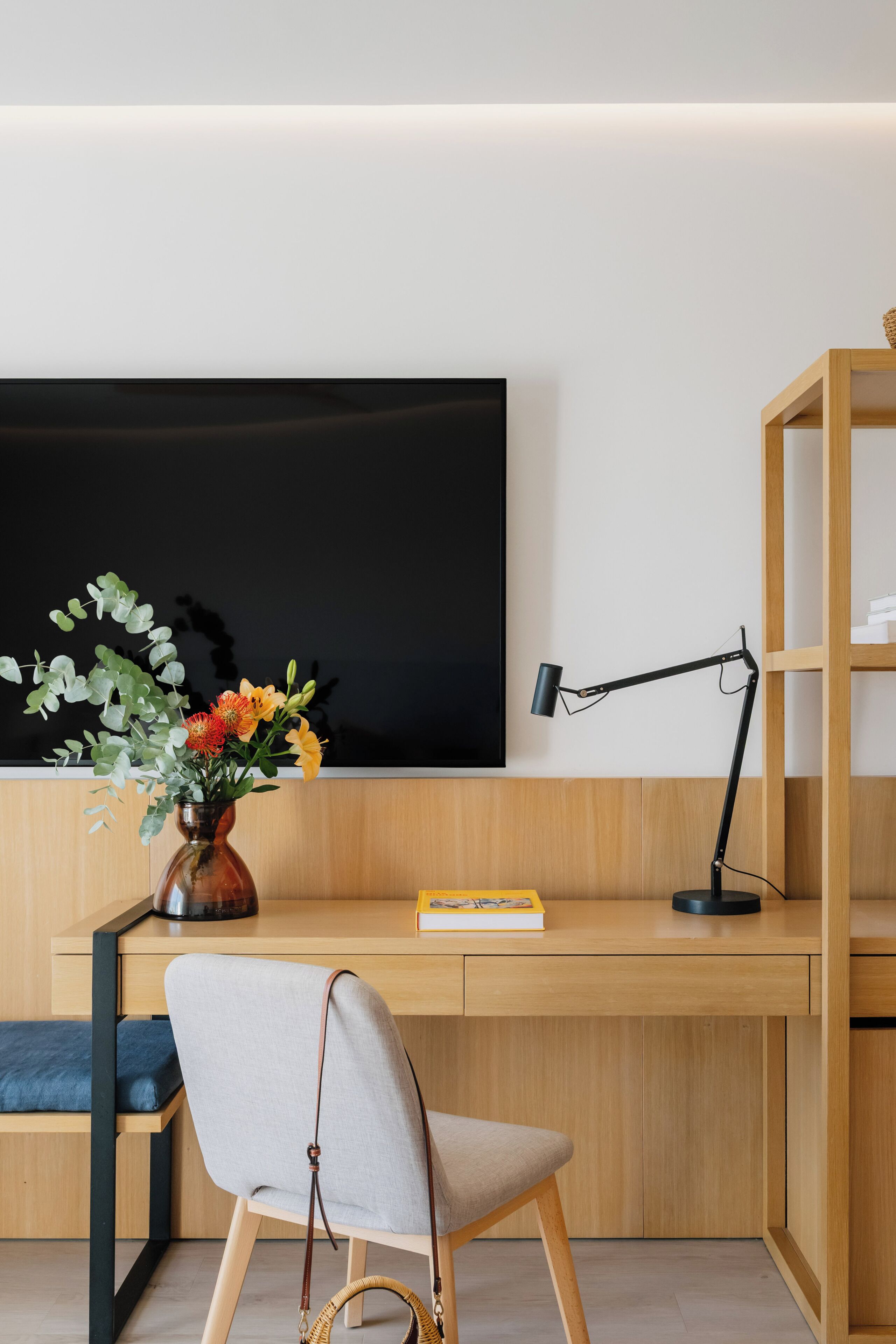 A modern workspace with a desk, chair, TV, lamp, bookshelf, and a vase with flowers.