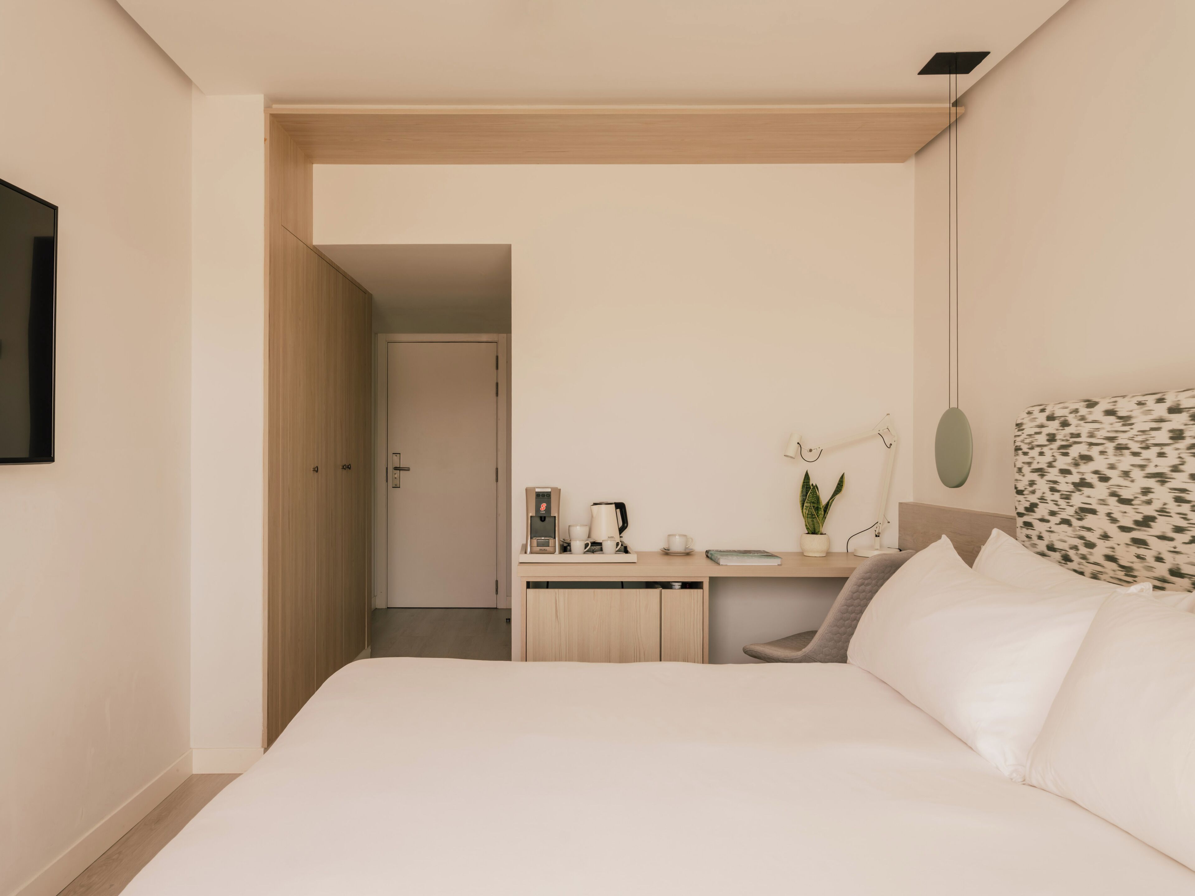 A bedroom with a bed, a desk with a coffee machine, a chair, and a plant.